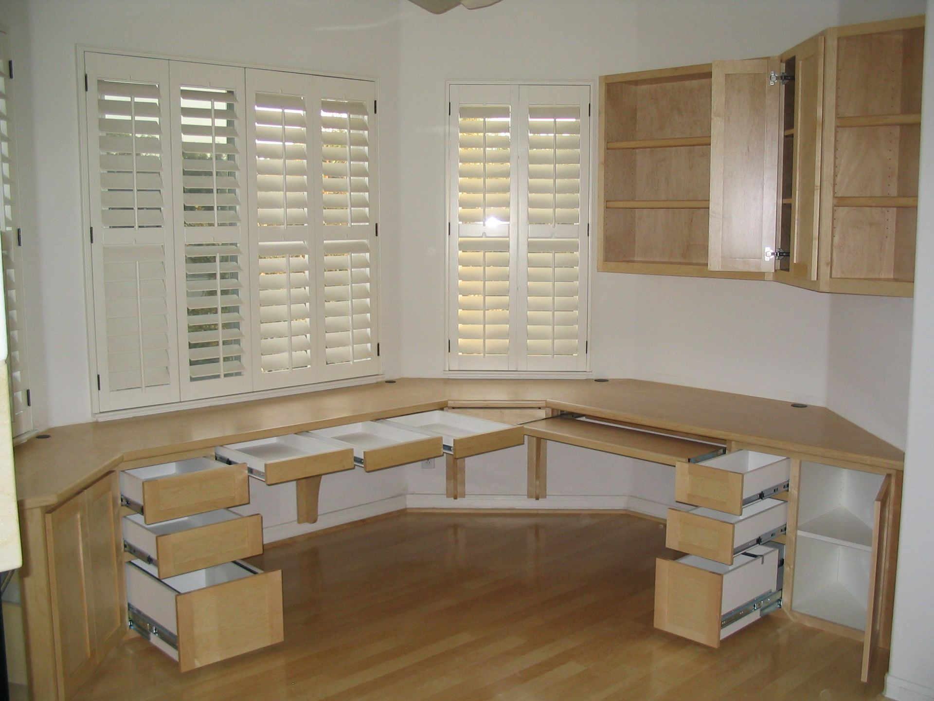 Built-in wooden desk in a corner room with two shuttered windows and upper cabinets; drawers are open.