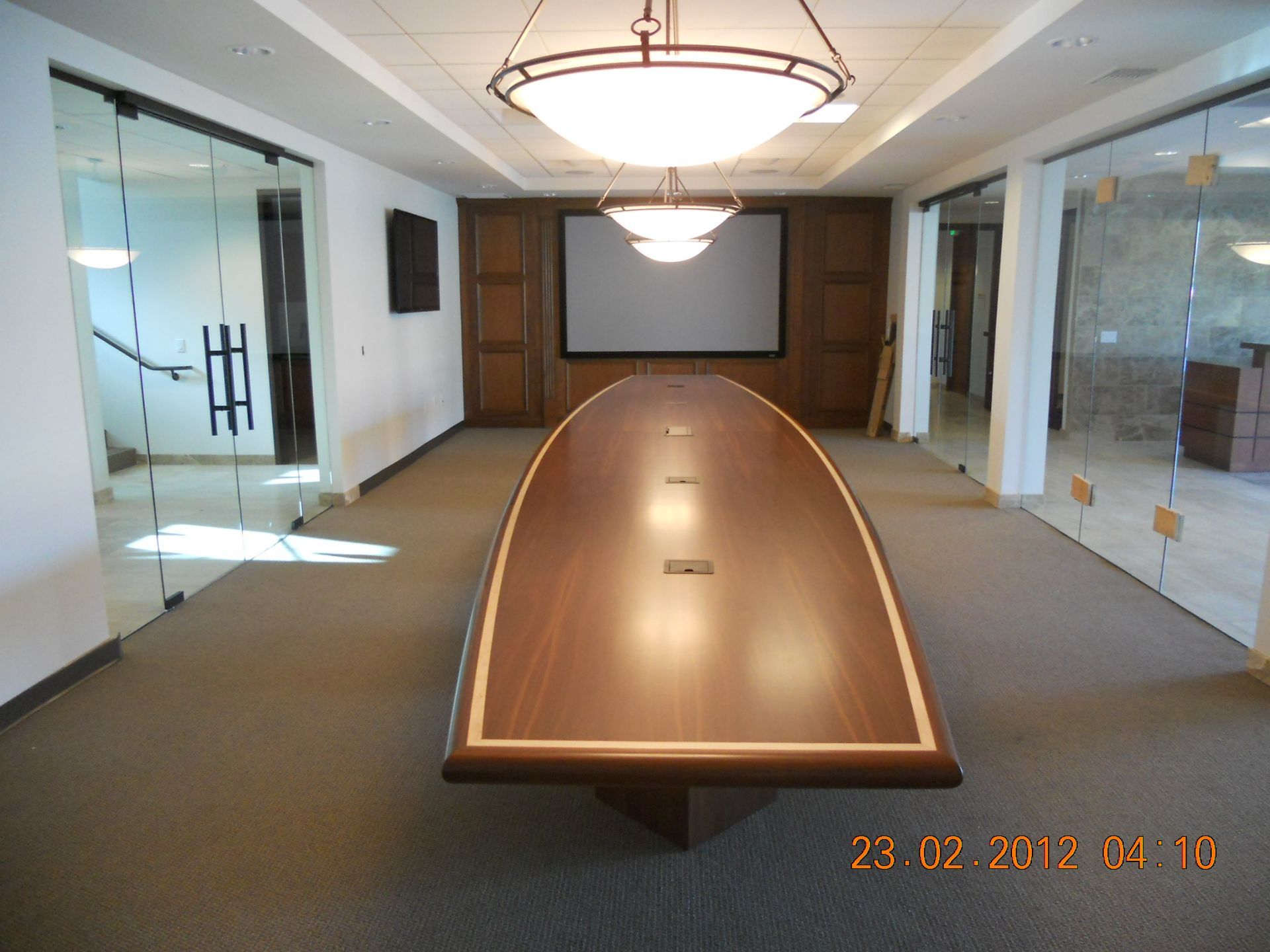 Conference room with large wooden table, projector screen, glass walls, and hanging lights.
