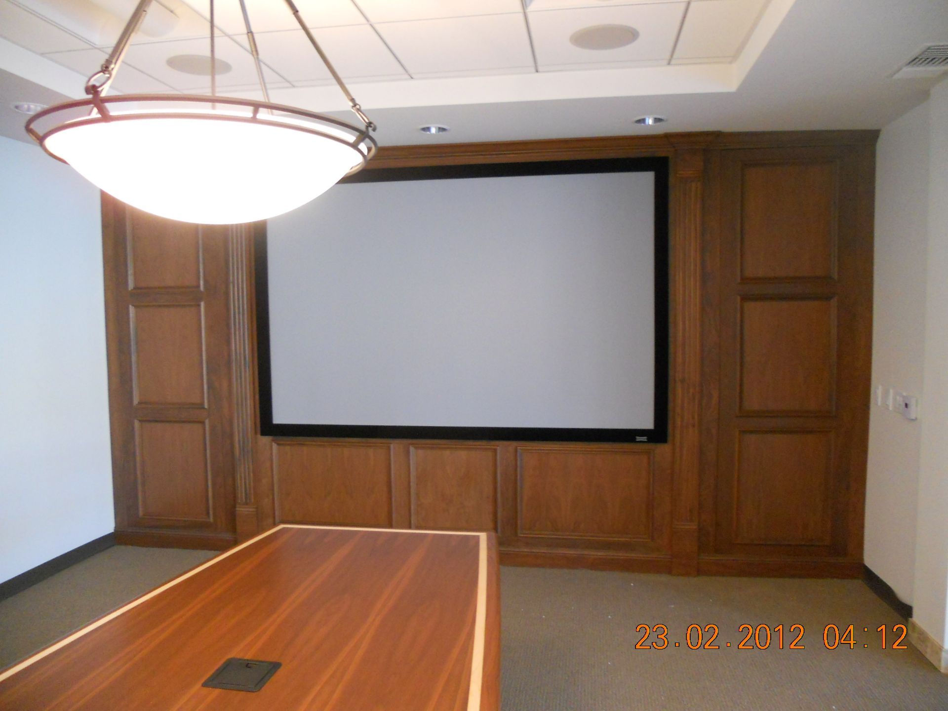 Conference room with wooden paneling, a large screen, and a table. Overhead light.