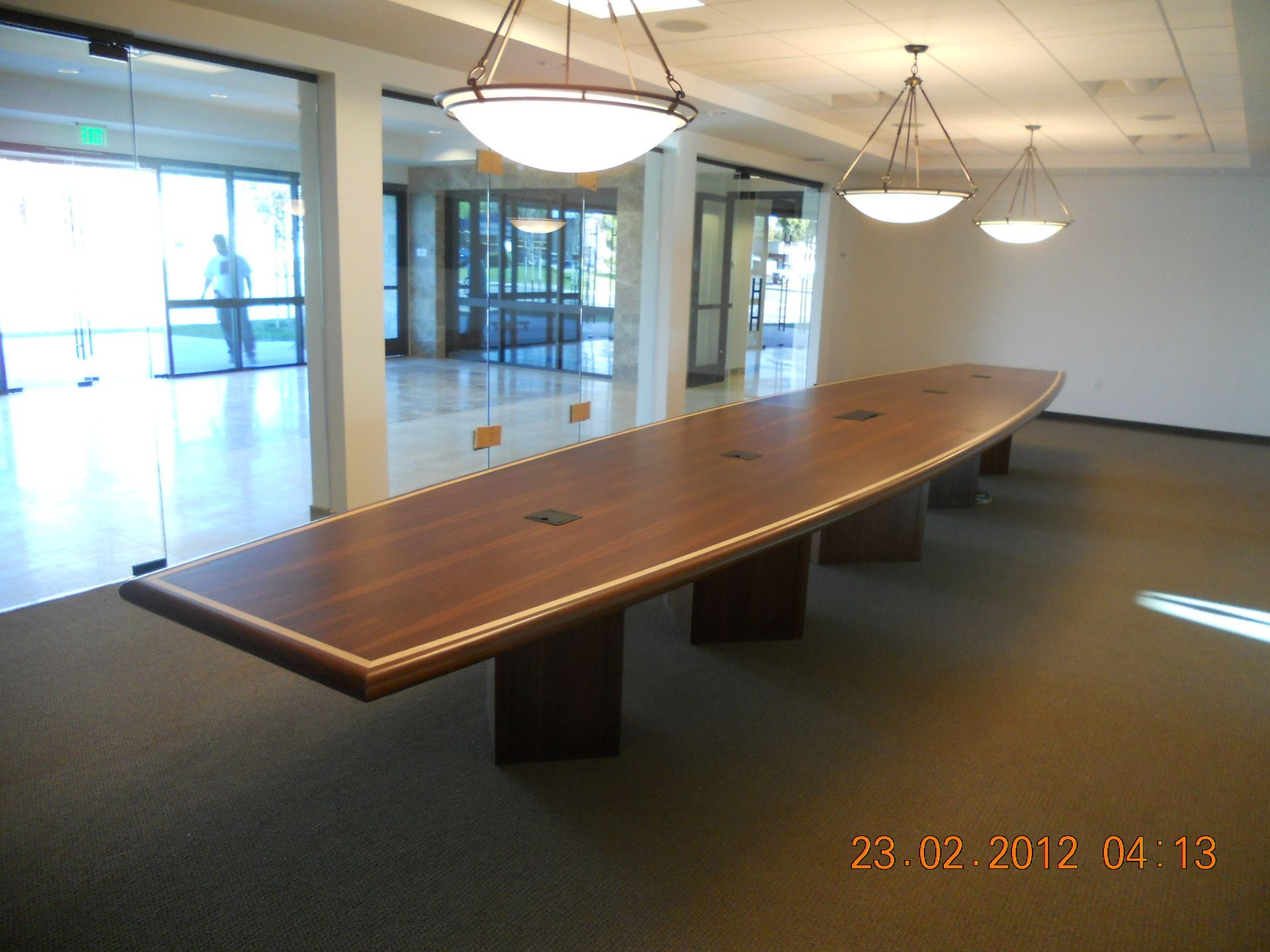 Long, wood conference table in a modern room with glass doors and hanging lights.