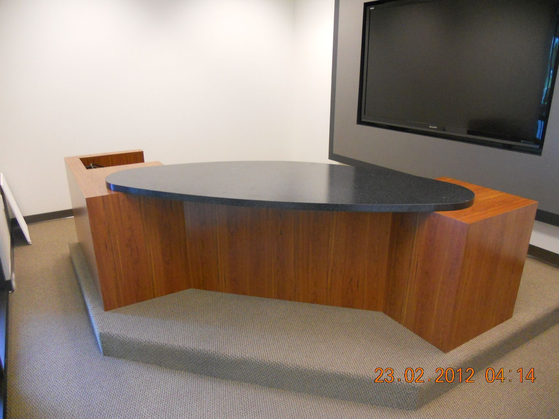 Modern wooden conference table with a dark top, against a neutral wall with a large TV screen.