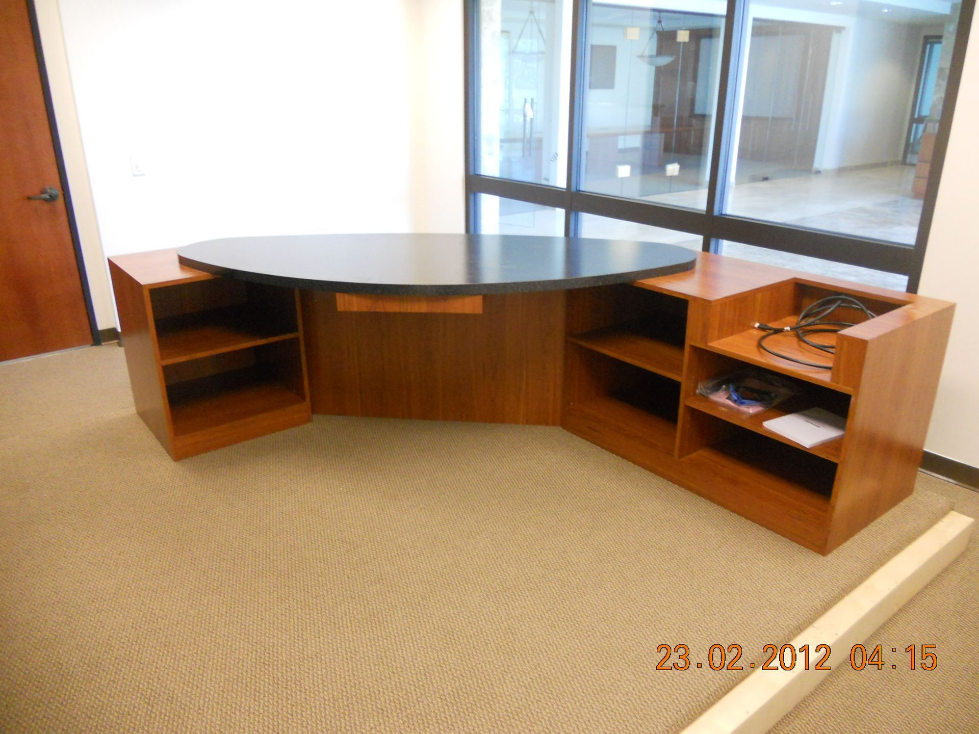Wooden reception desk with a black countertop and built-in shelving on beige carpet.