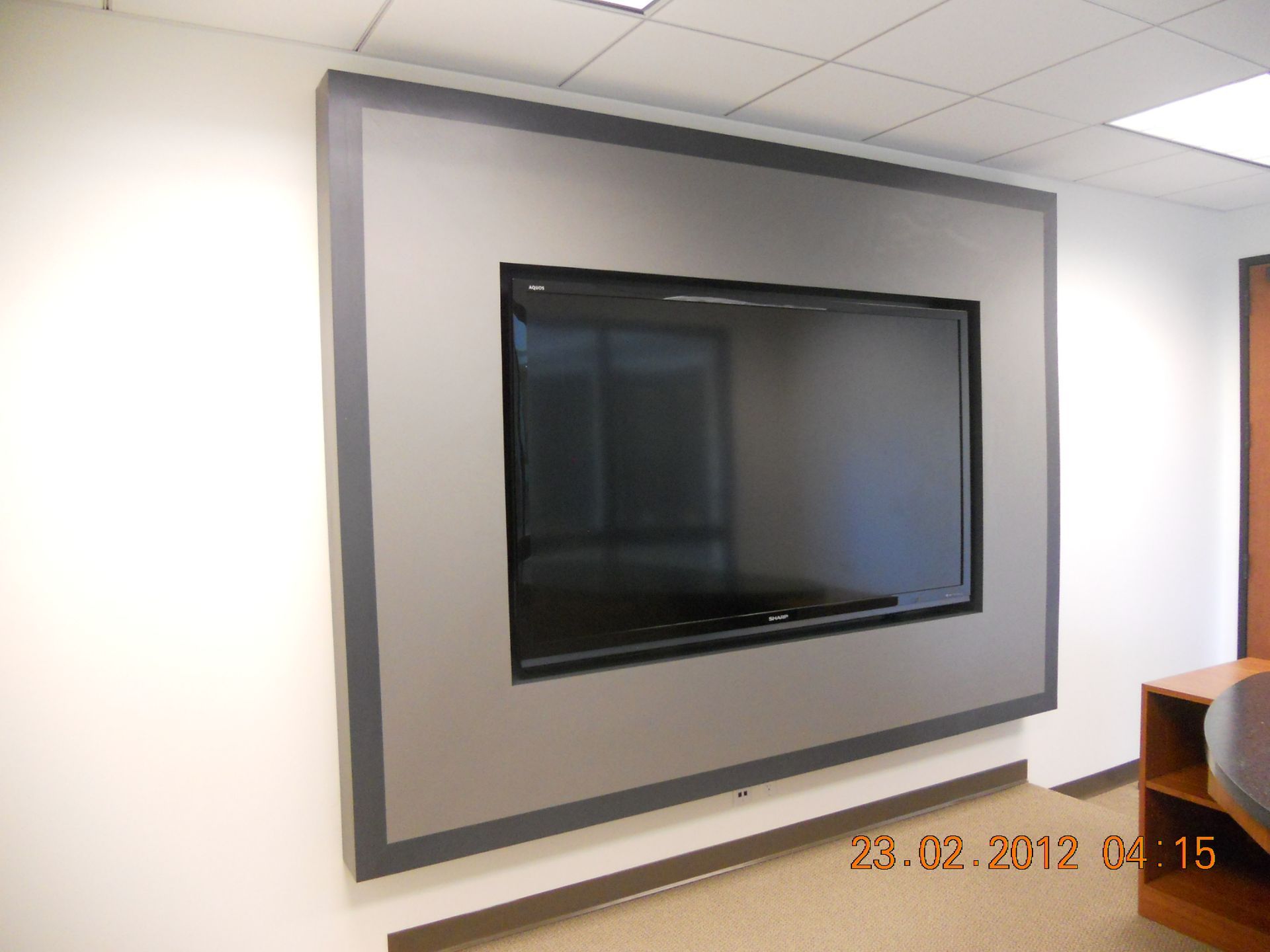 Large flat-screen TV mounted on a wall with a gray border. The TV reflects an office setting.