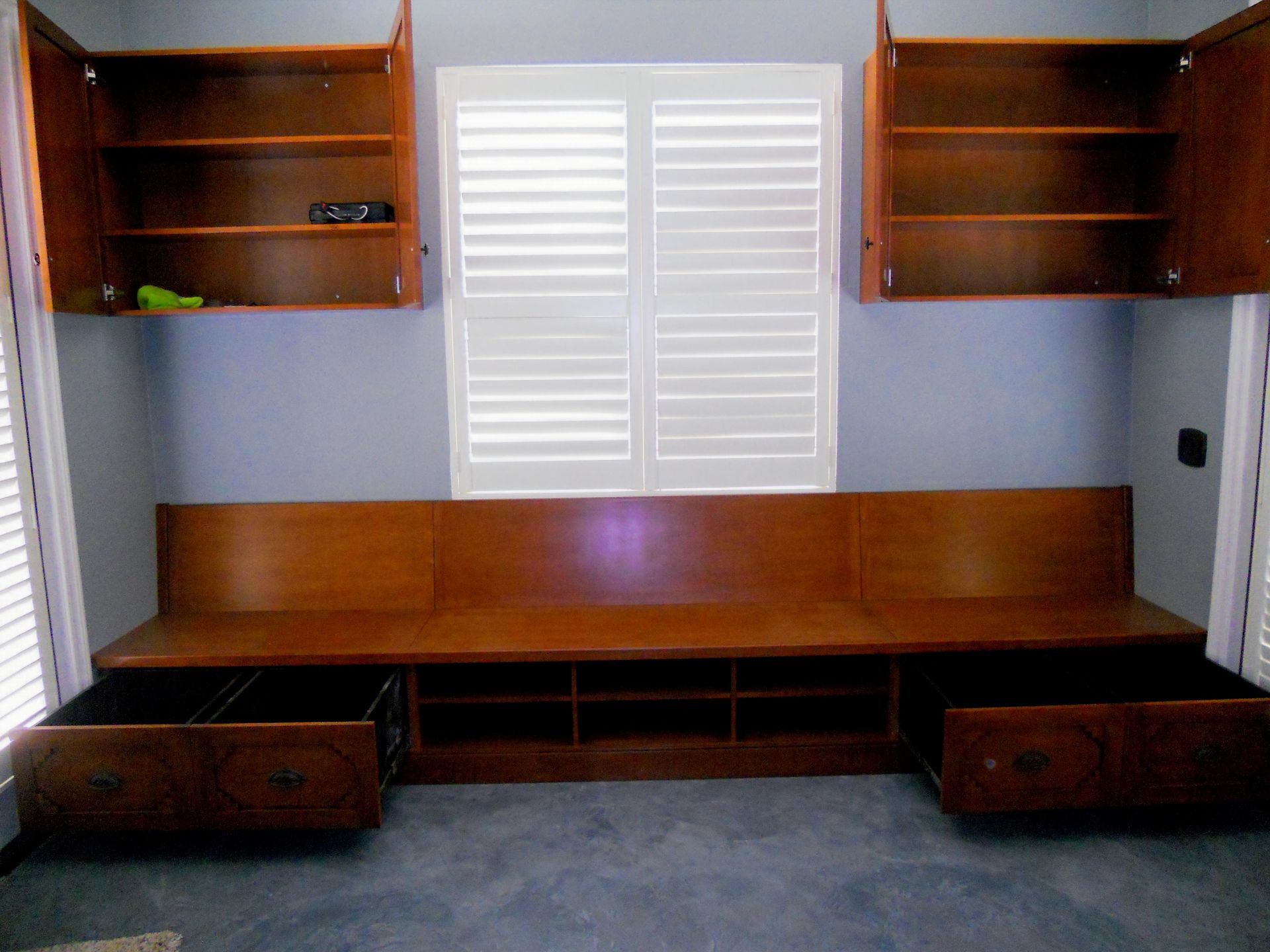 Wooden storage bench with open drawers and shelves, built-in beneath a window with open cabinets above.