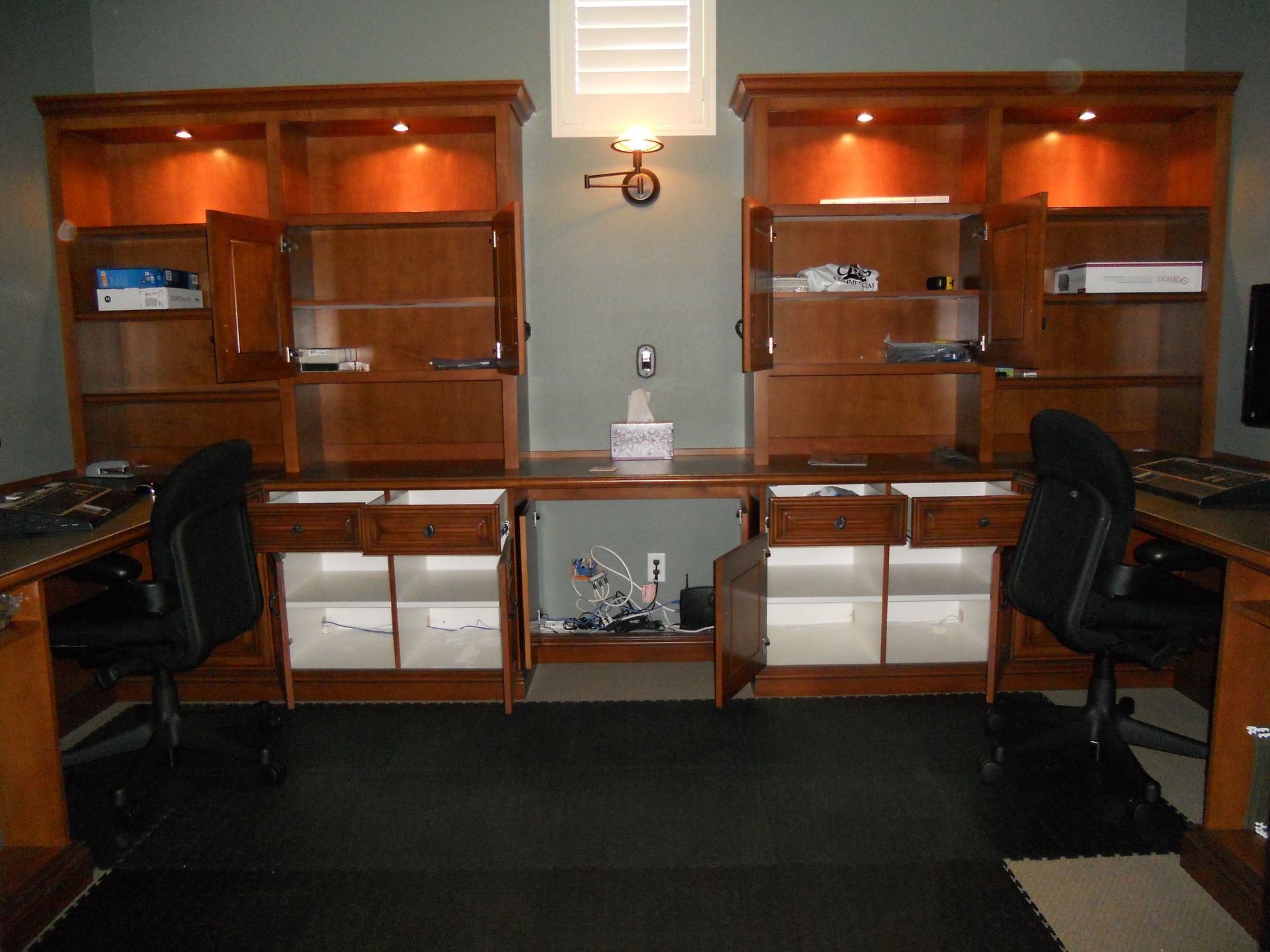 Two wooden office desks with open cabinets and built-in shelving, two black chairs, and a light-colored wall.