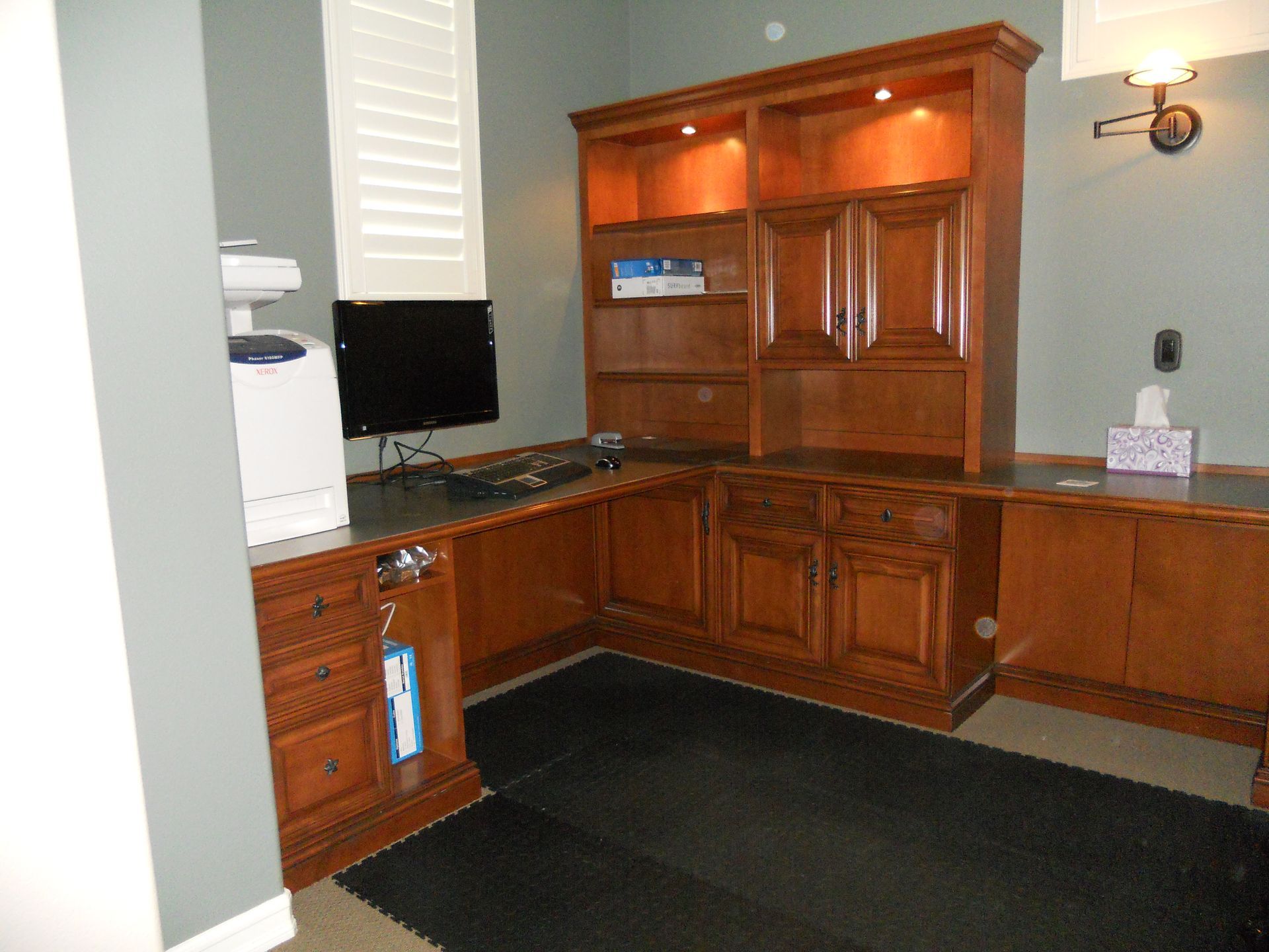 Wooden office desk with a computer monitor and built-in bookshelves. Green wall and black floor mat.