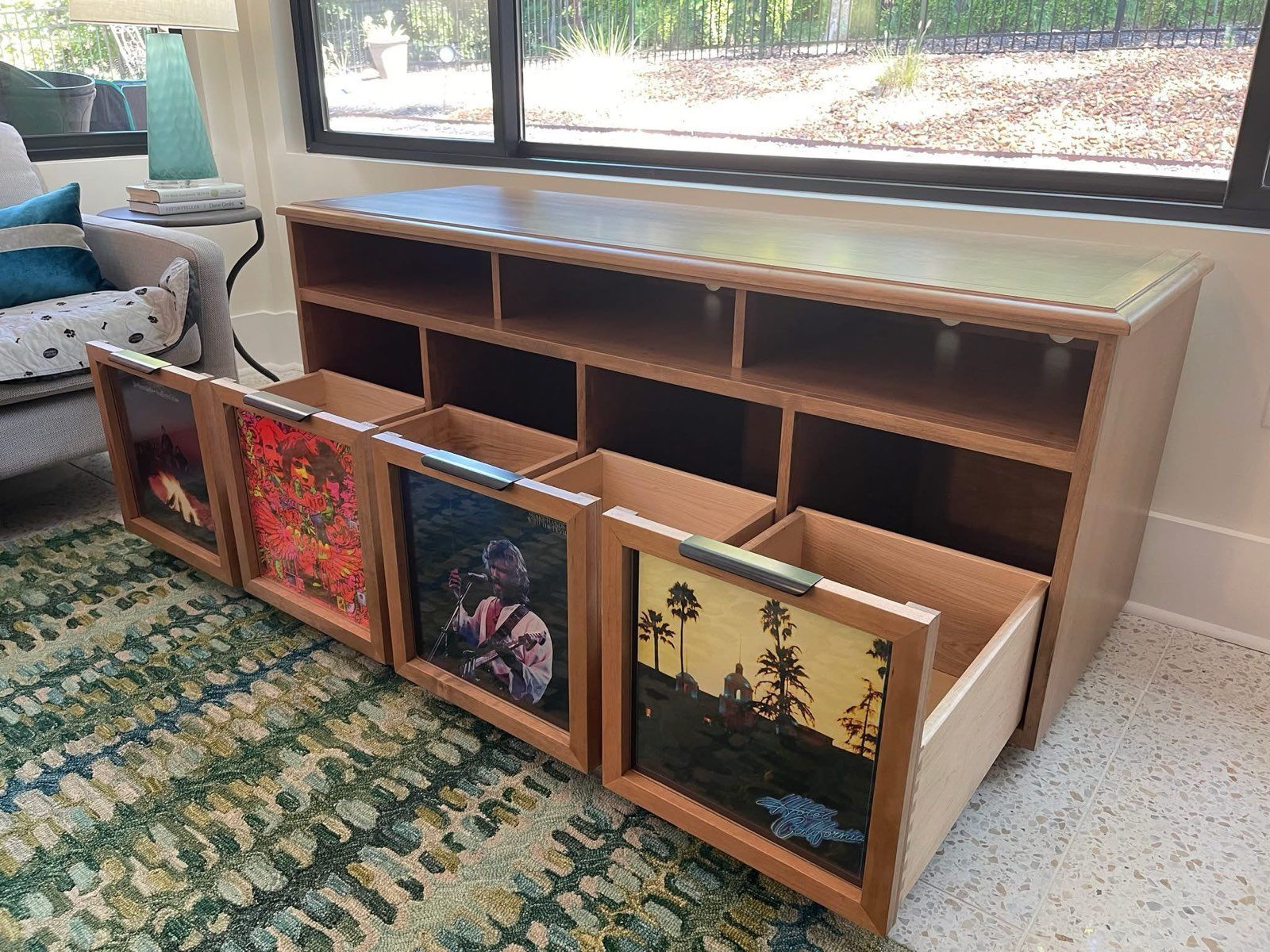 Wooden record storage cabinet with framed album art on the drawers, near a window and rug.