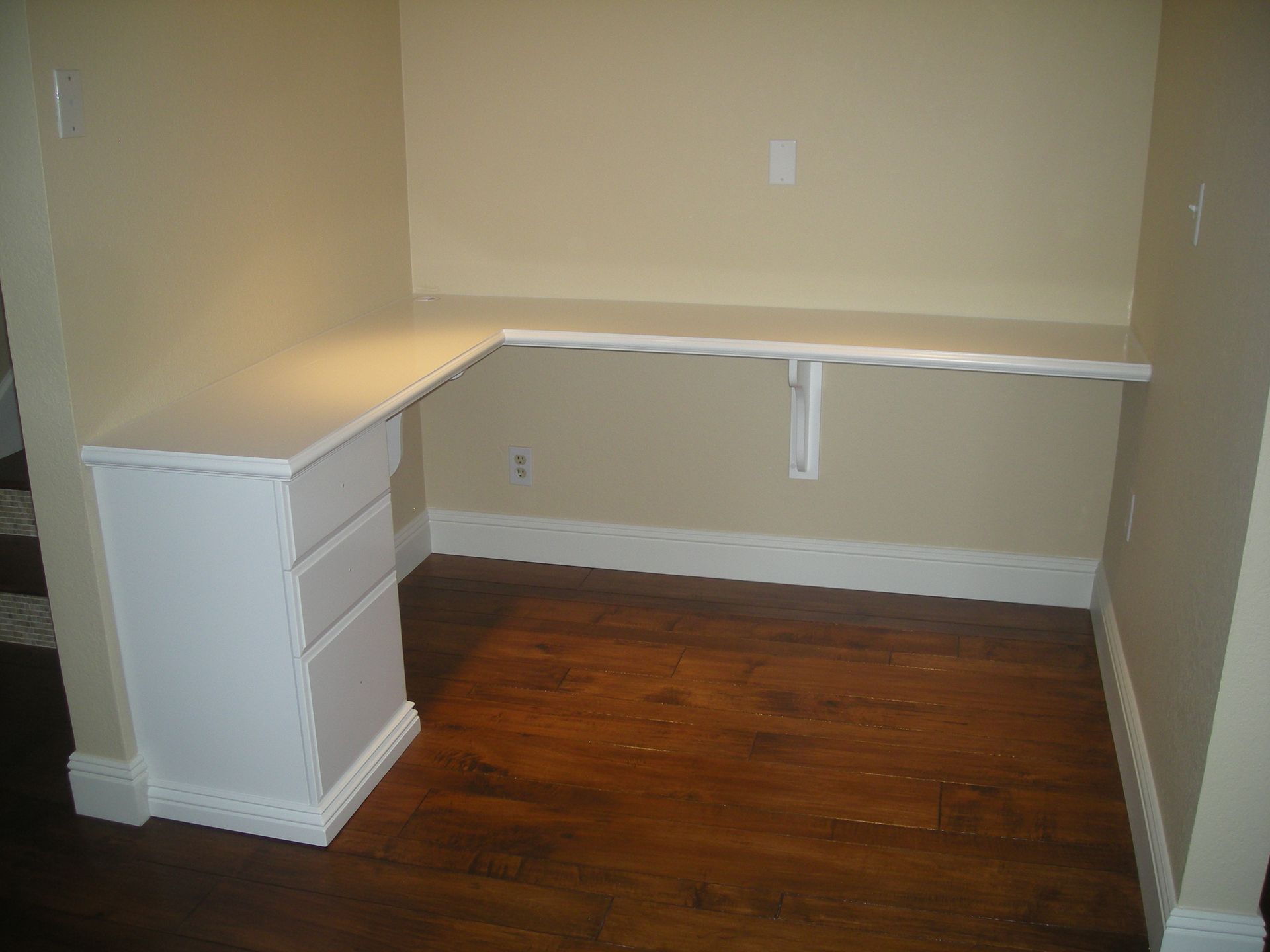 Built-in white desk with drawers and shelf, against beige walls and wood floor.