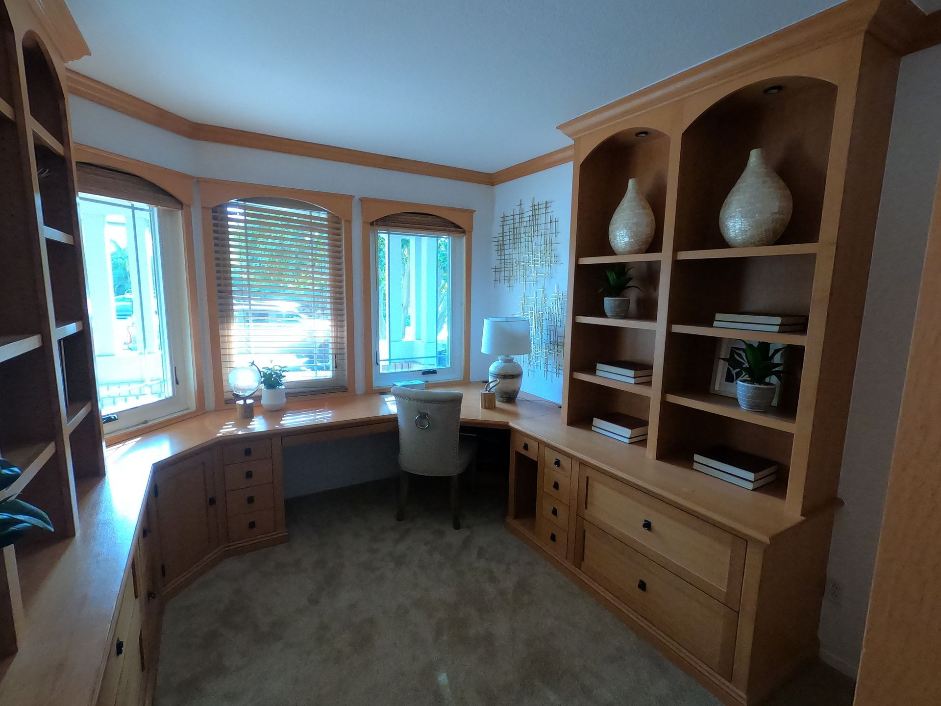 Home office with wooden desk, bookshelves, bay window with blinds. Beige walls and carpet.