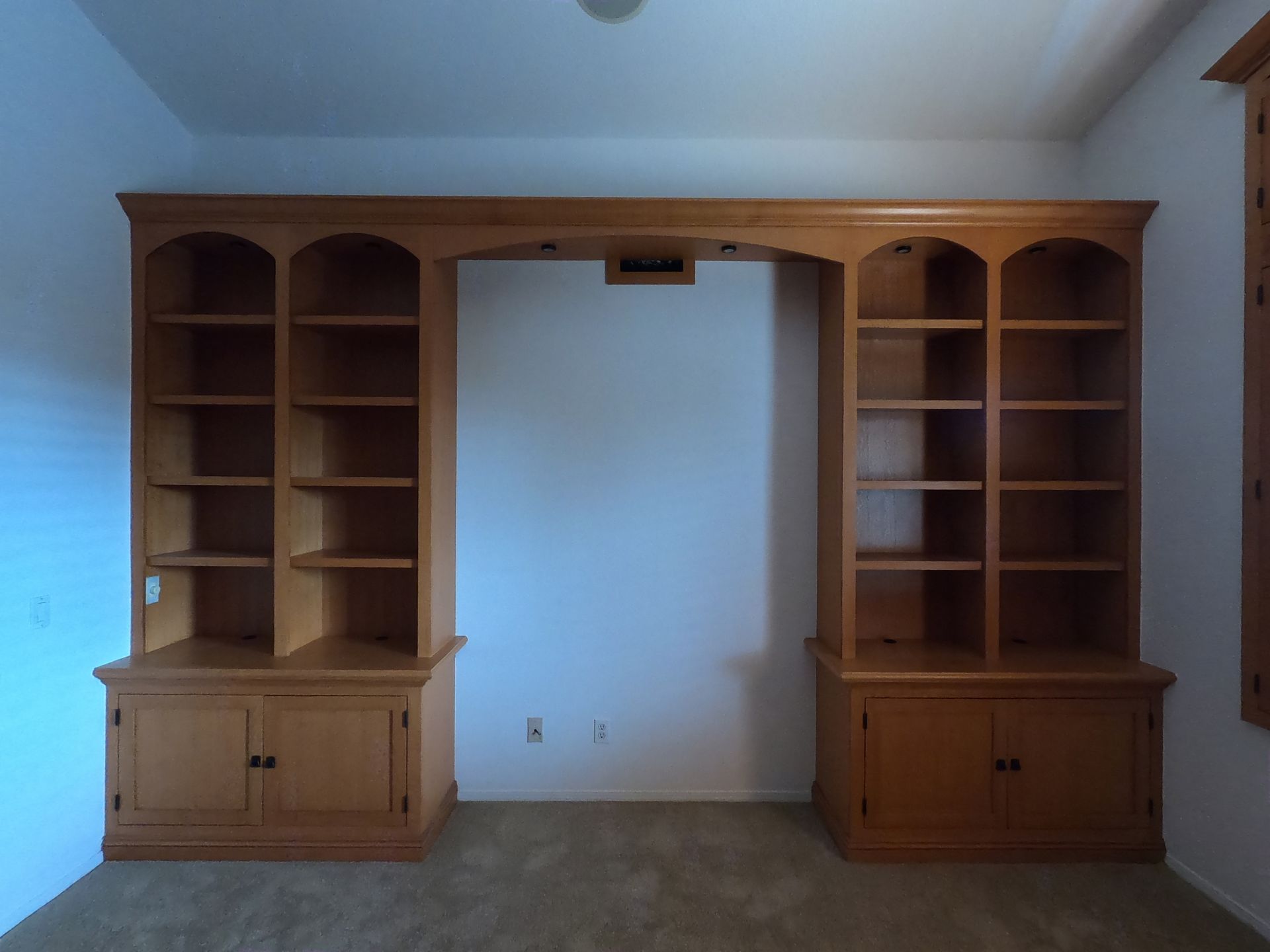 Built-in wooden bookshelves on either side of a blank wall. Brown cabinets below with arch-topped shelves above.