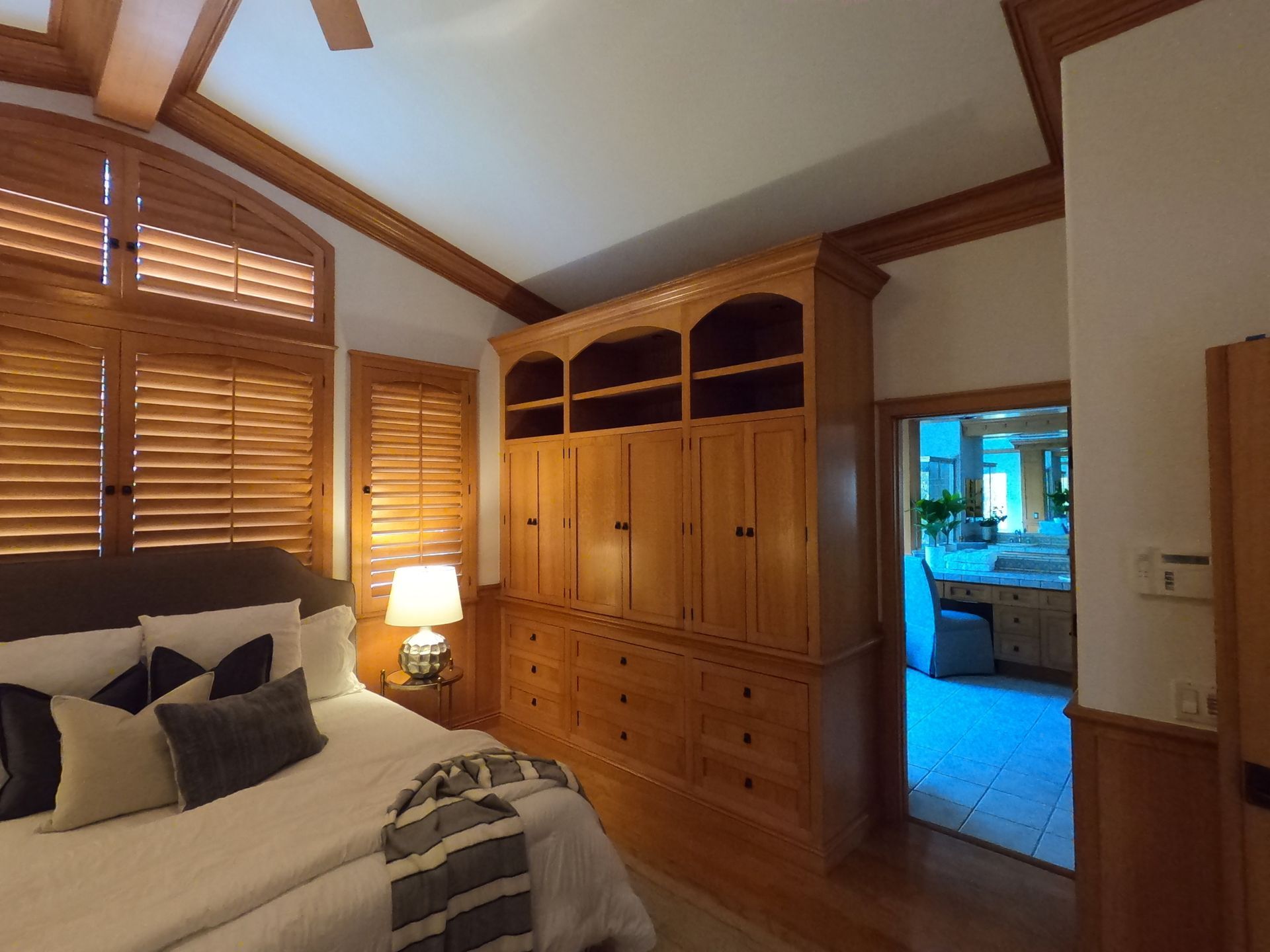 Bedroom with wooden built-in storage, bed with pillows, open doorway to another room, and shuttered windows.
