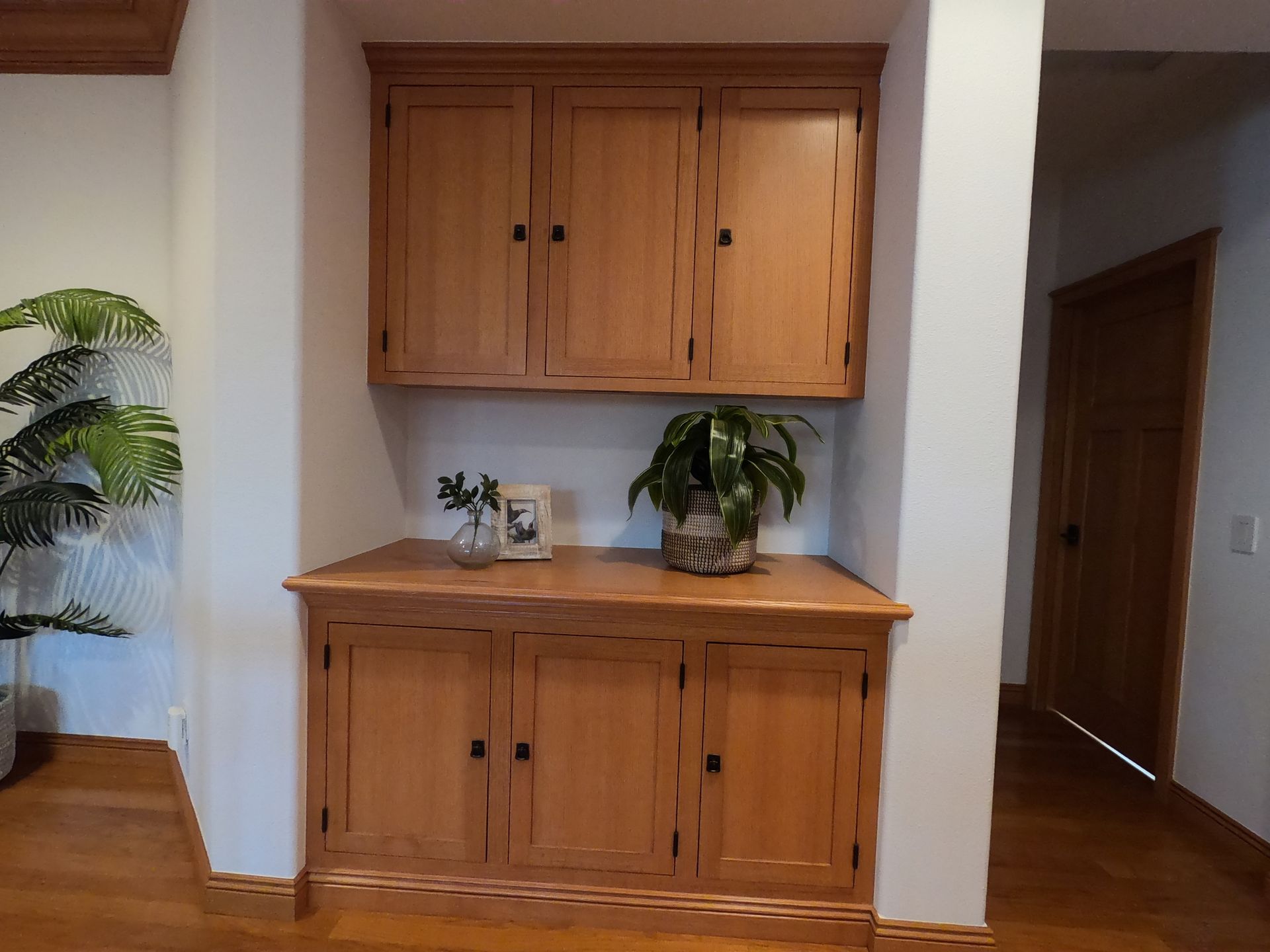 Wooden built-in cabinet with upper and lower sections, open shelf with plants, in an alcove with a doorway.