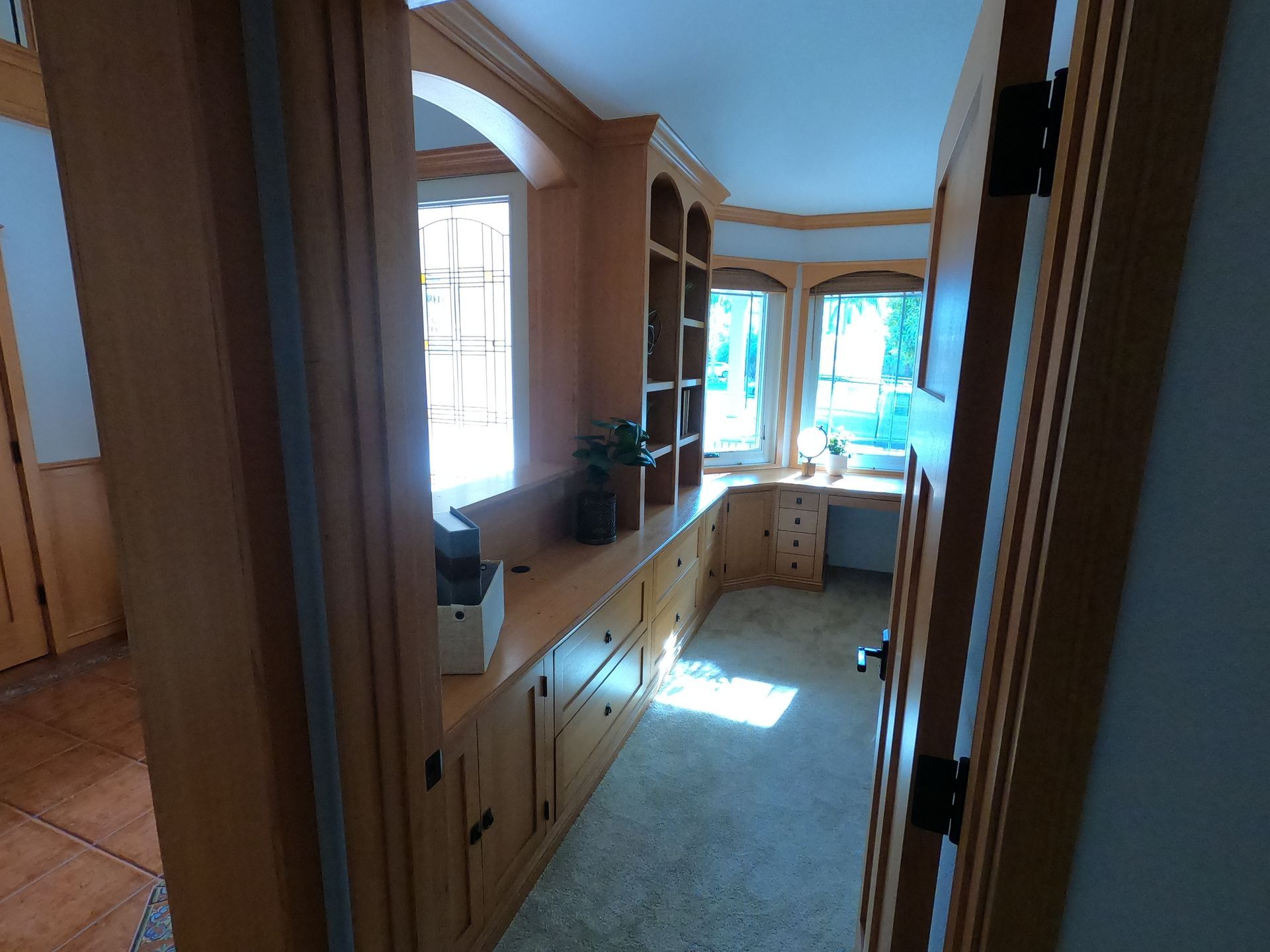 Wooden built-in office with arched windows, cabinets, and desk. Room has natural light and a beige carpet.