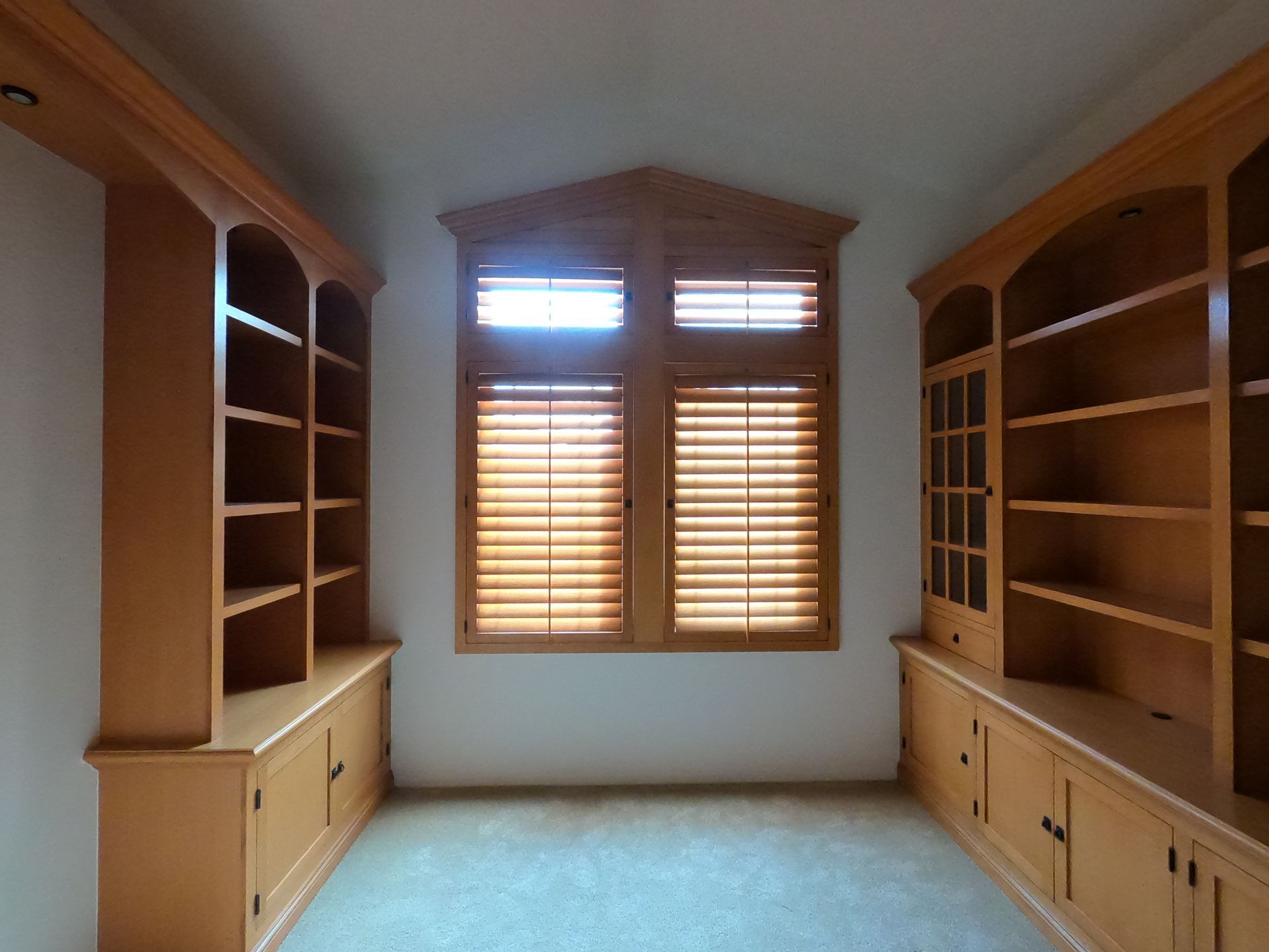 Built-in wooden bookshelves and cabinets flank a window with blinds; room with beige carpet.