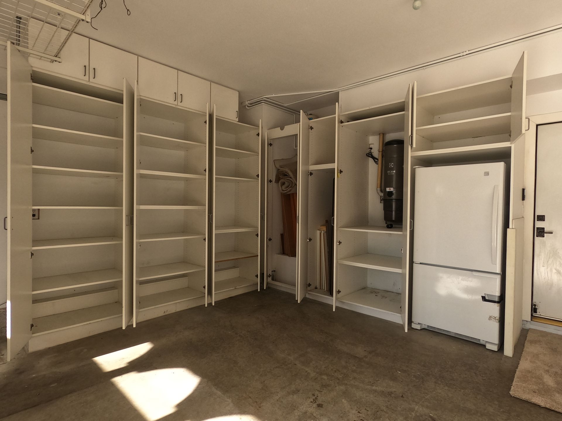 Garage interior with white cabinets and shelves, a refrigerator, and a concrete floor.