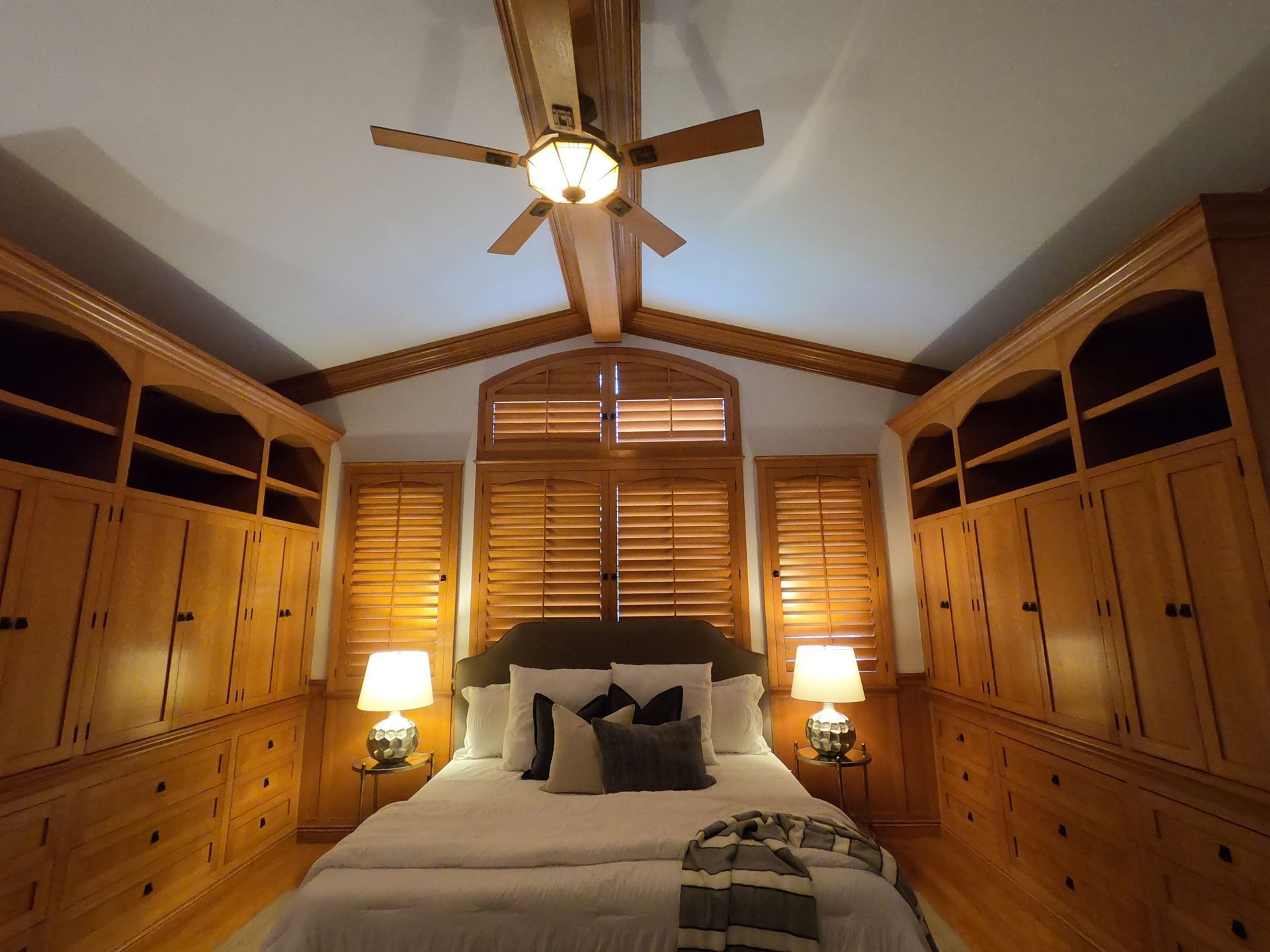 Bedroom with wooden built-ins, shutters, and a ceiling fan over a bed.