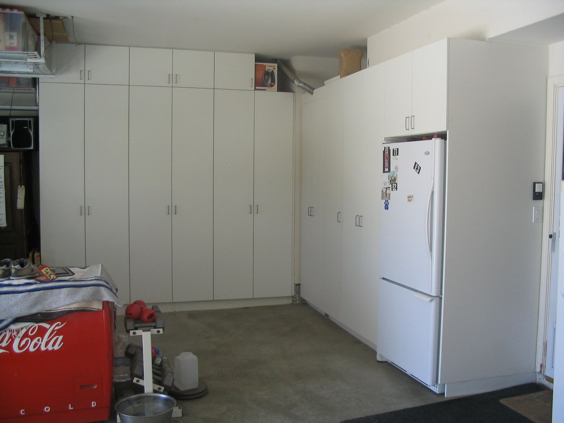 Garage interior with white cabinets and a refrigerator. A vintage Coca-Cola cooler and other items sit on the floor.