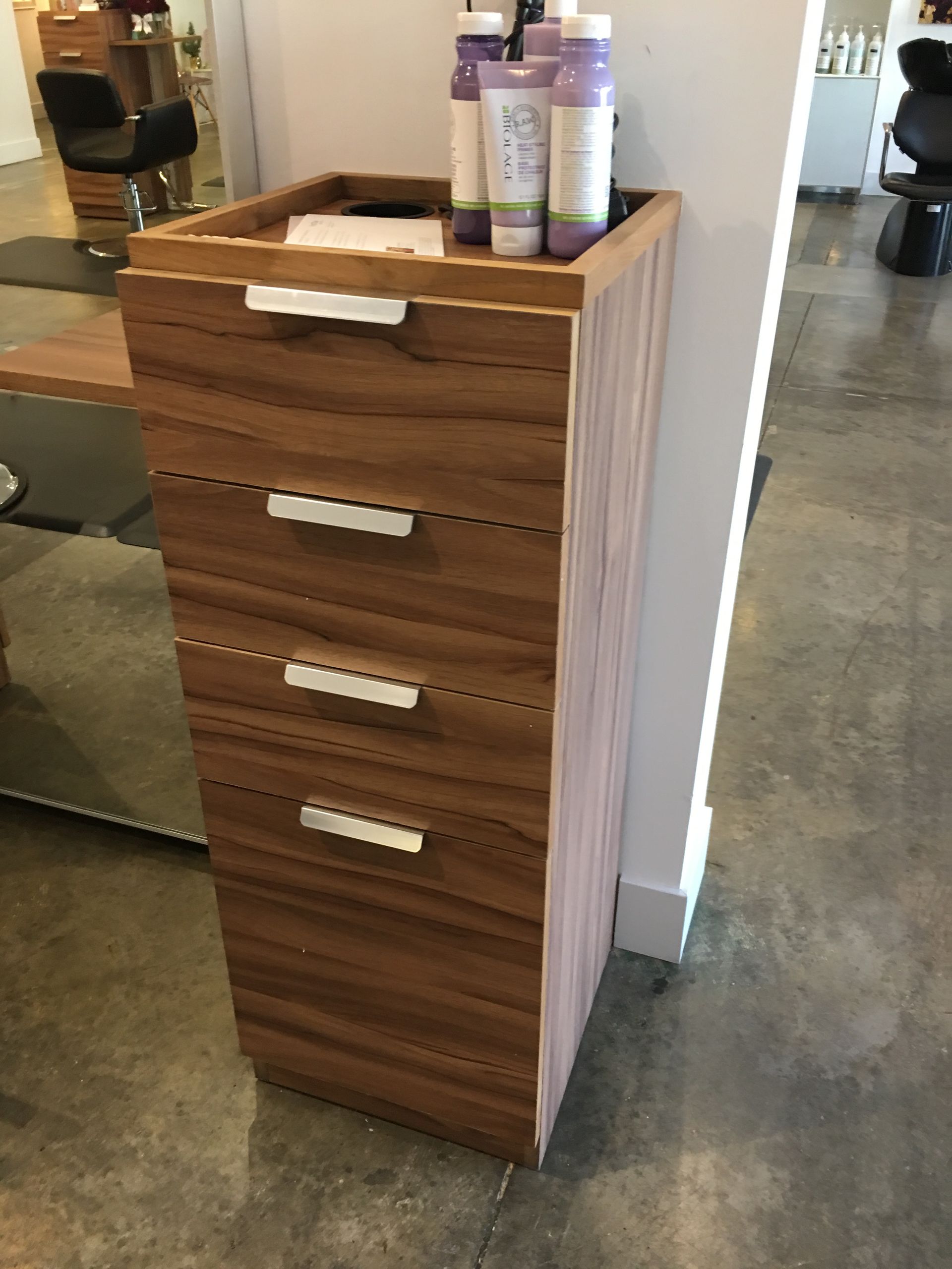 Brown wooden cabinet with four drawers, silver handles, and products on top, next to a white wall.