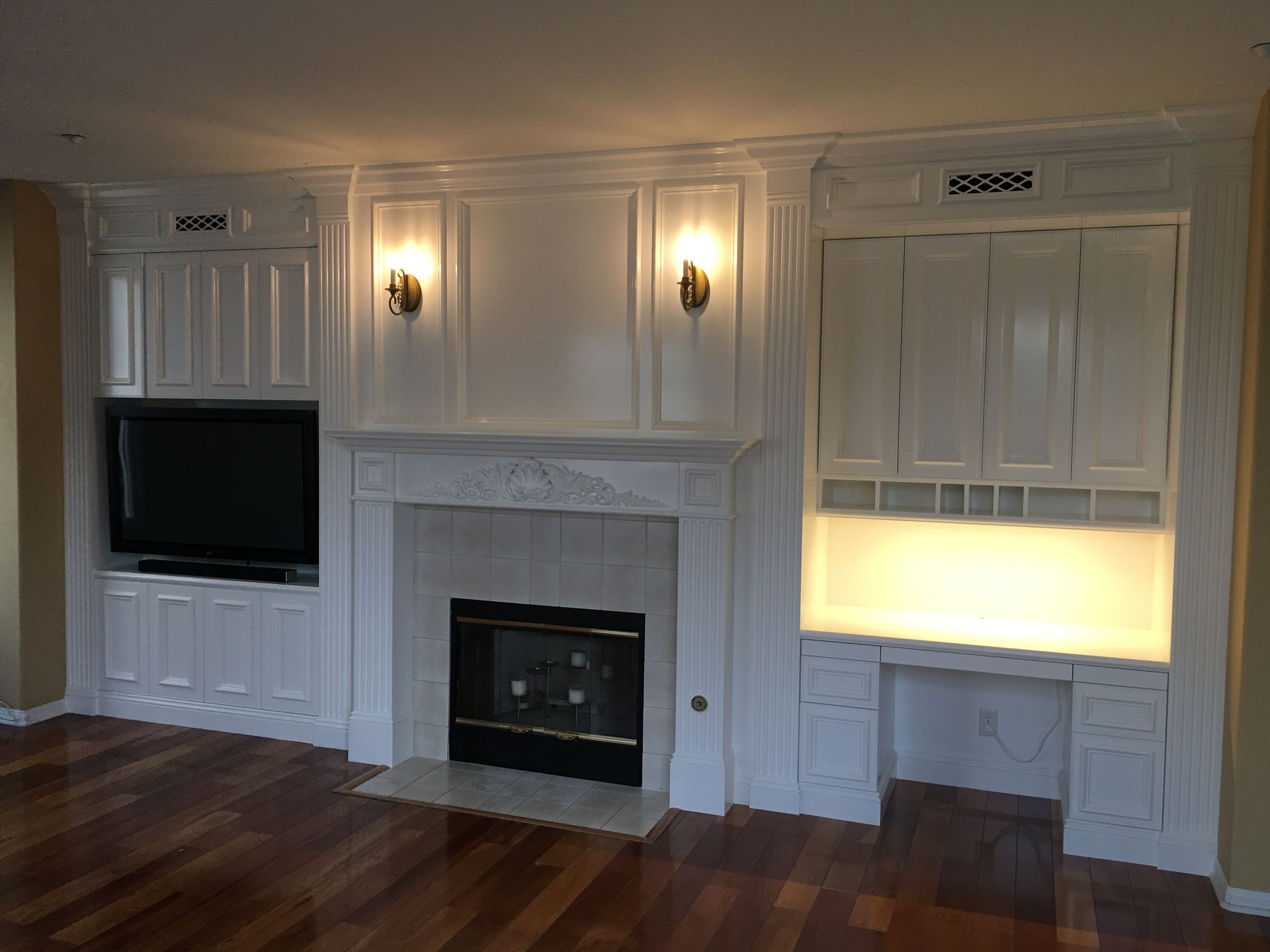 White built-in fireplace with cabinets and a TV on a wood floor. Two sconces flank the mantel.