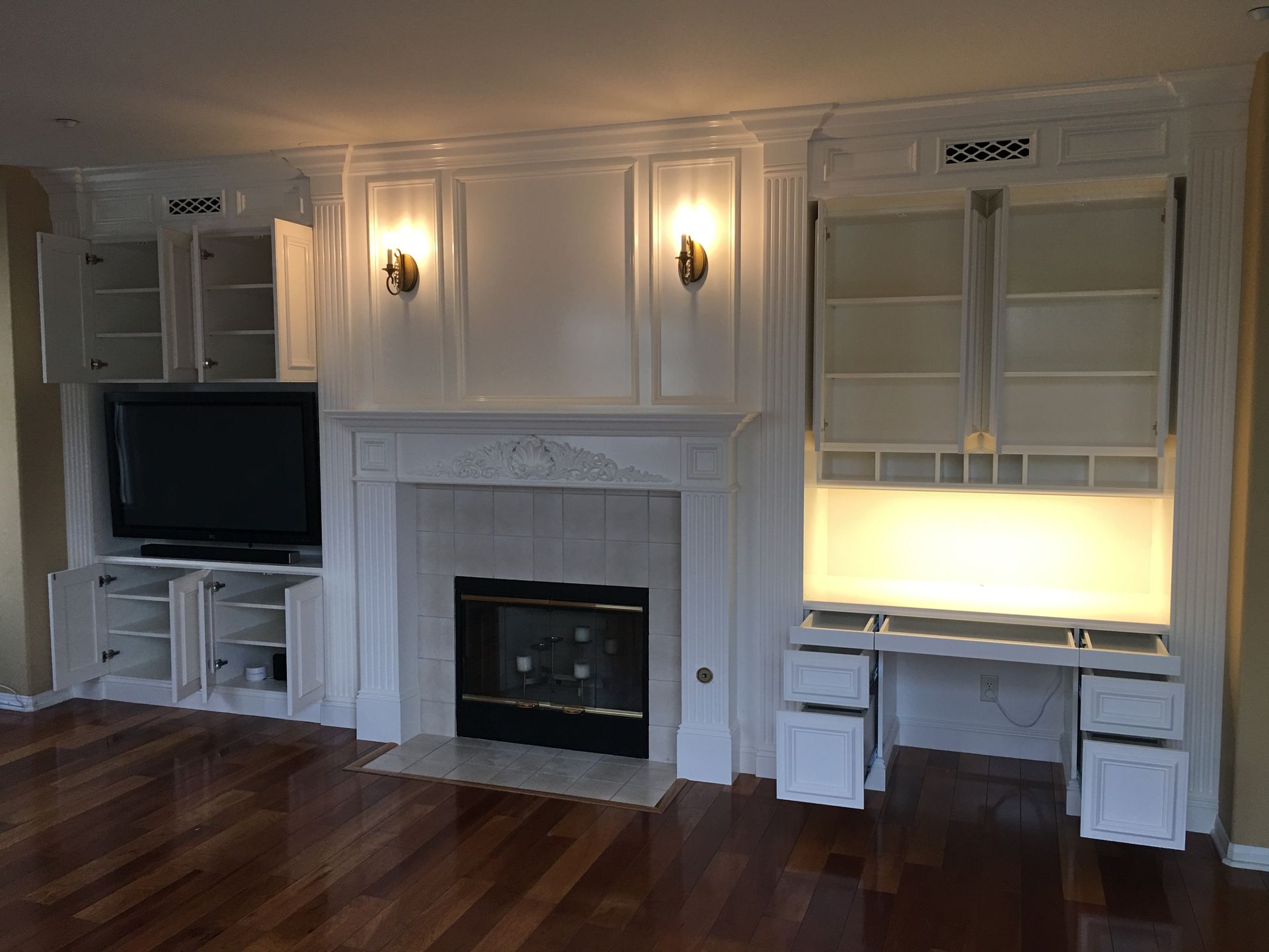 Built-in white cabinets flanking a fireplace with a black TV, desk, and sconces in a room with wood floors.