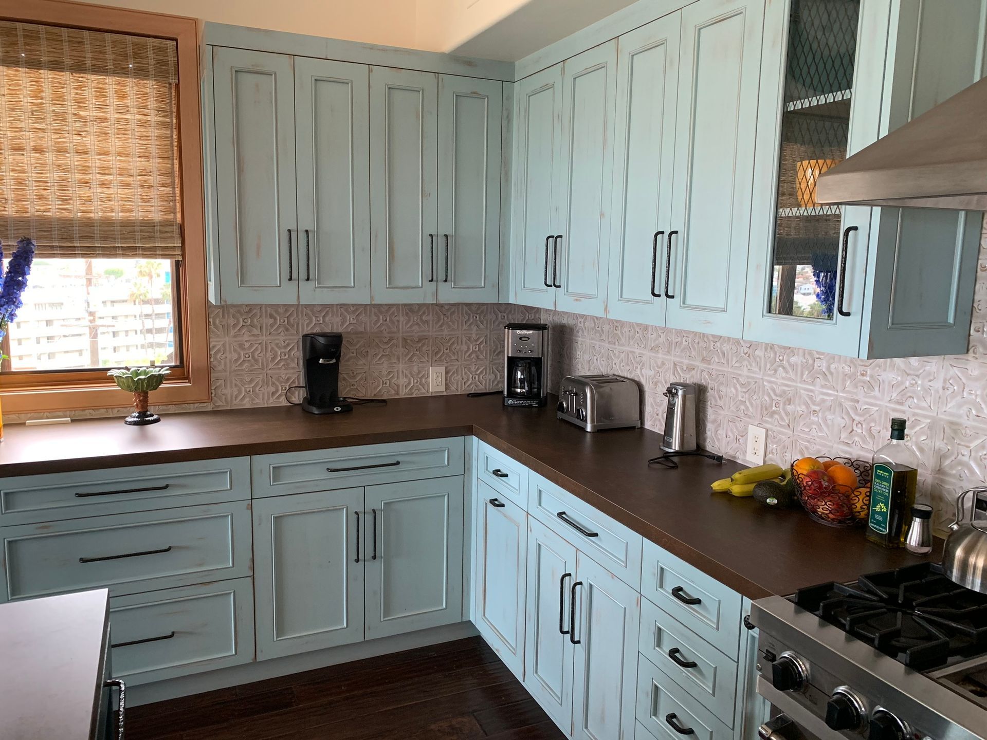 Blue kitchen cabinets with dark countertops, stainless steel appliances, and a window with a woven shade.