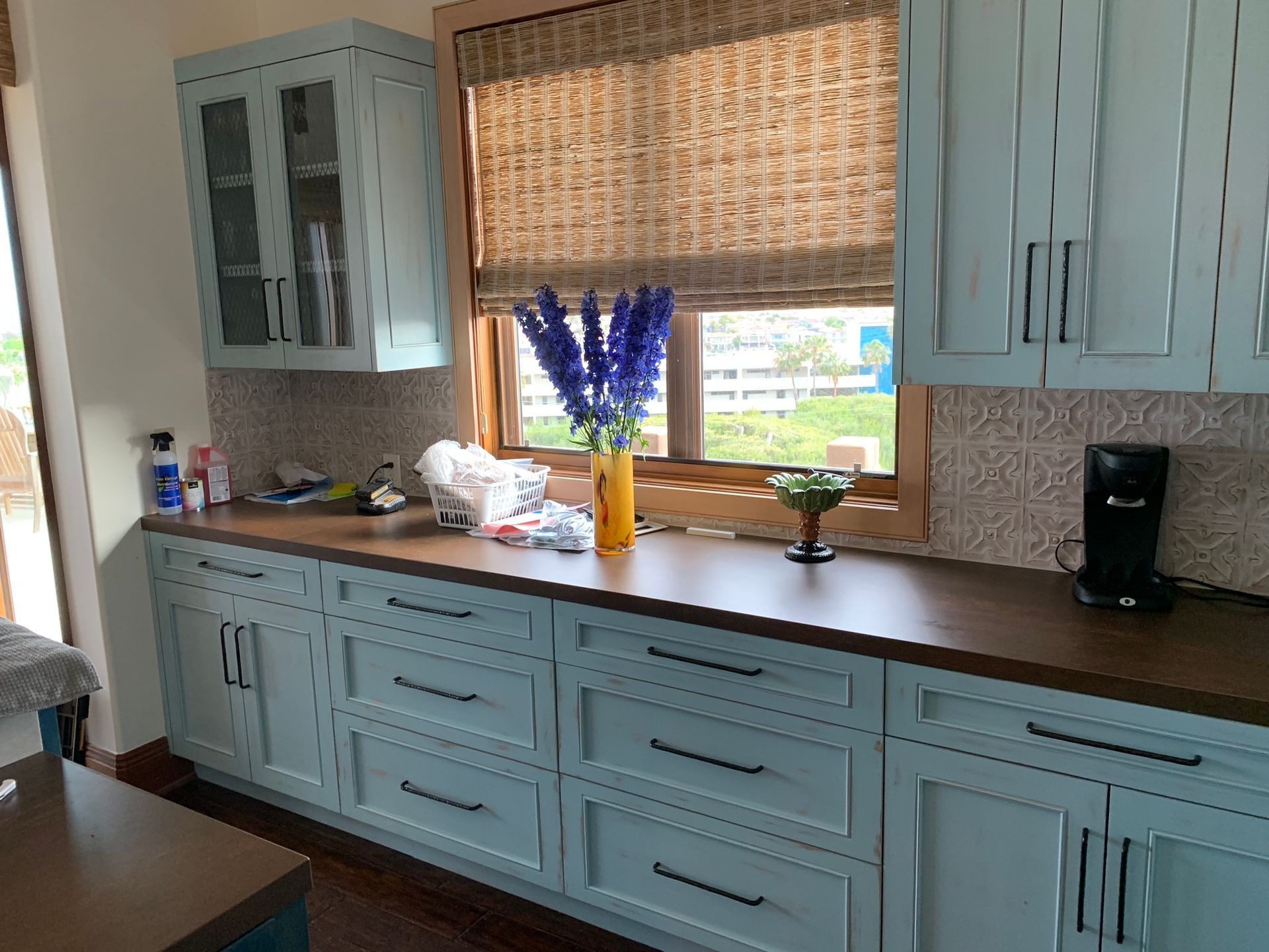 Light blue kitchen cabinets with a dark countertop and woven window shade.