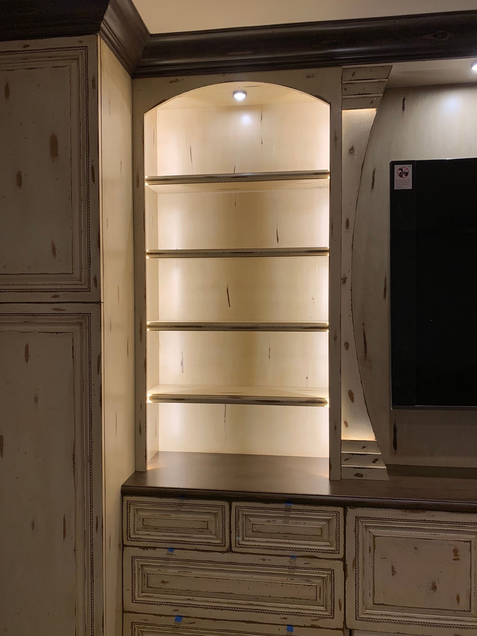 Built-in cabinet with arched shelves, illuminated from within. White-painted wood with distressed finish, dark trim.