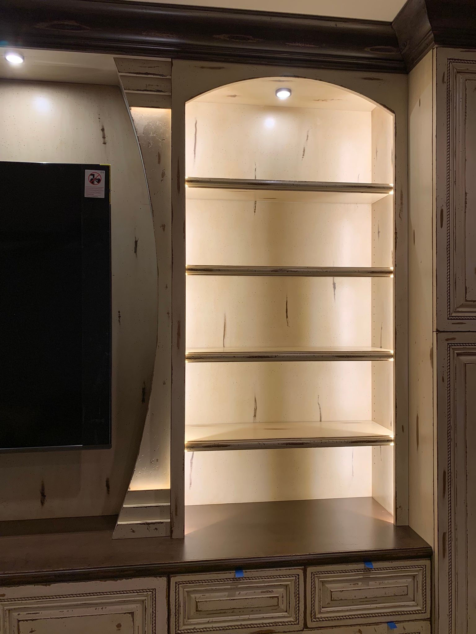Built-in cabinet with arched shelves and interior lighting. Dark wood trim and distressed cream finish.