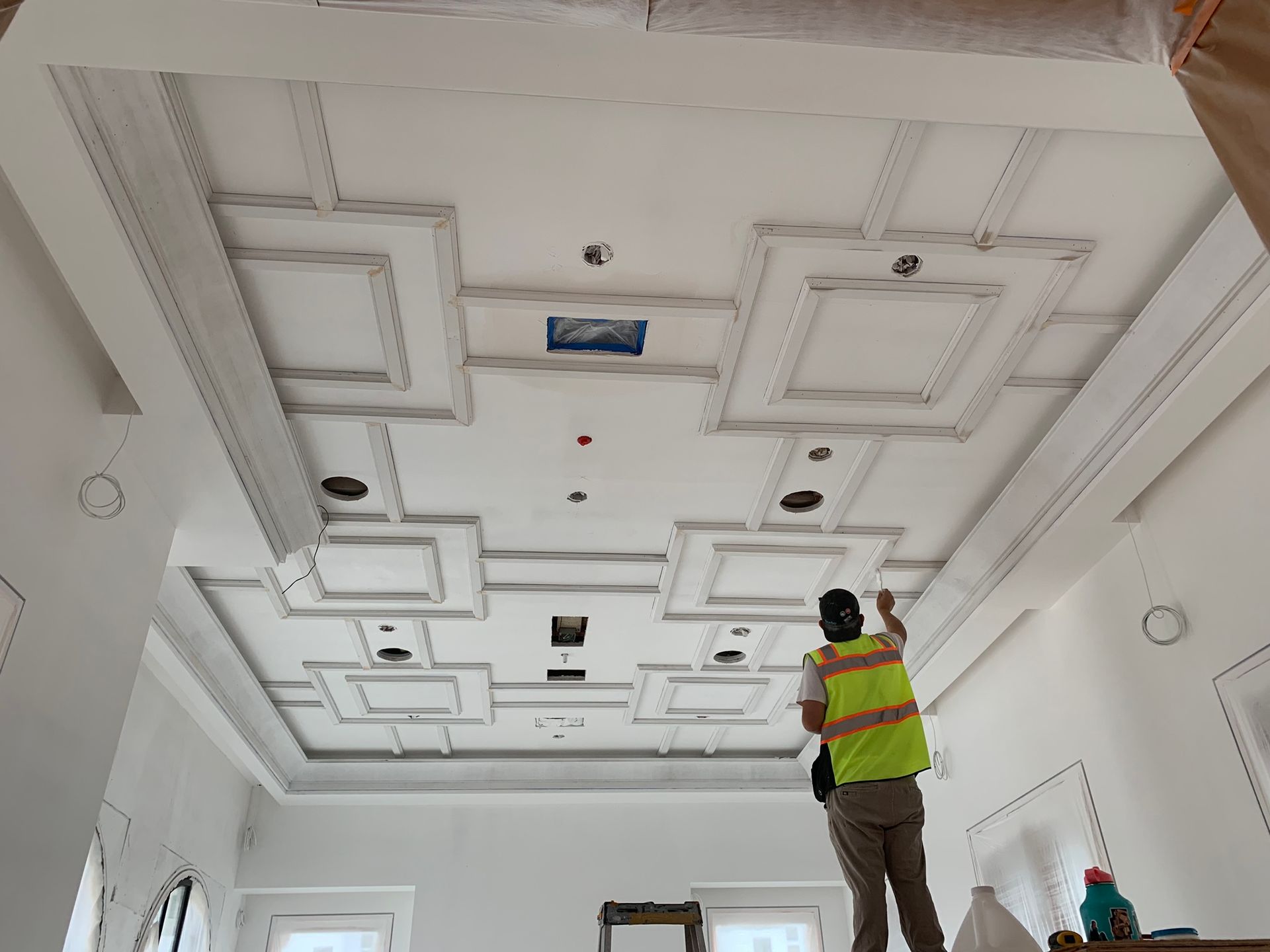 A person paints a white ornate ceiling with recessed lighting. They wear a safety vest, in a construction setting.