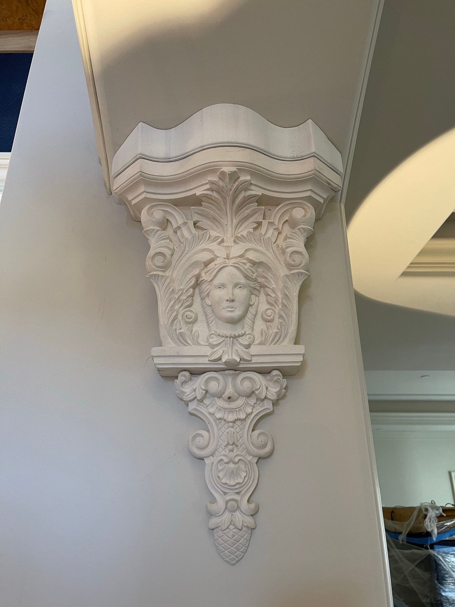 Ornate white plaster corbel with a woman's face sculpture on a light gray wall.