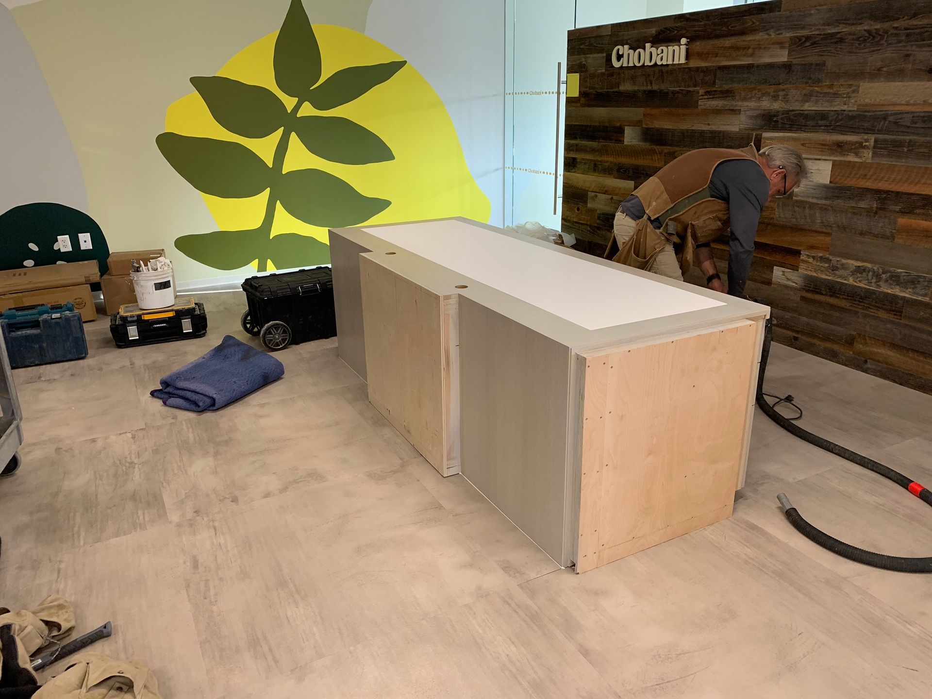 A carpenter is installing a countertop on a cabinet in an office. Wooden accent wall and leaf mural visible.