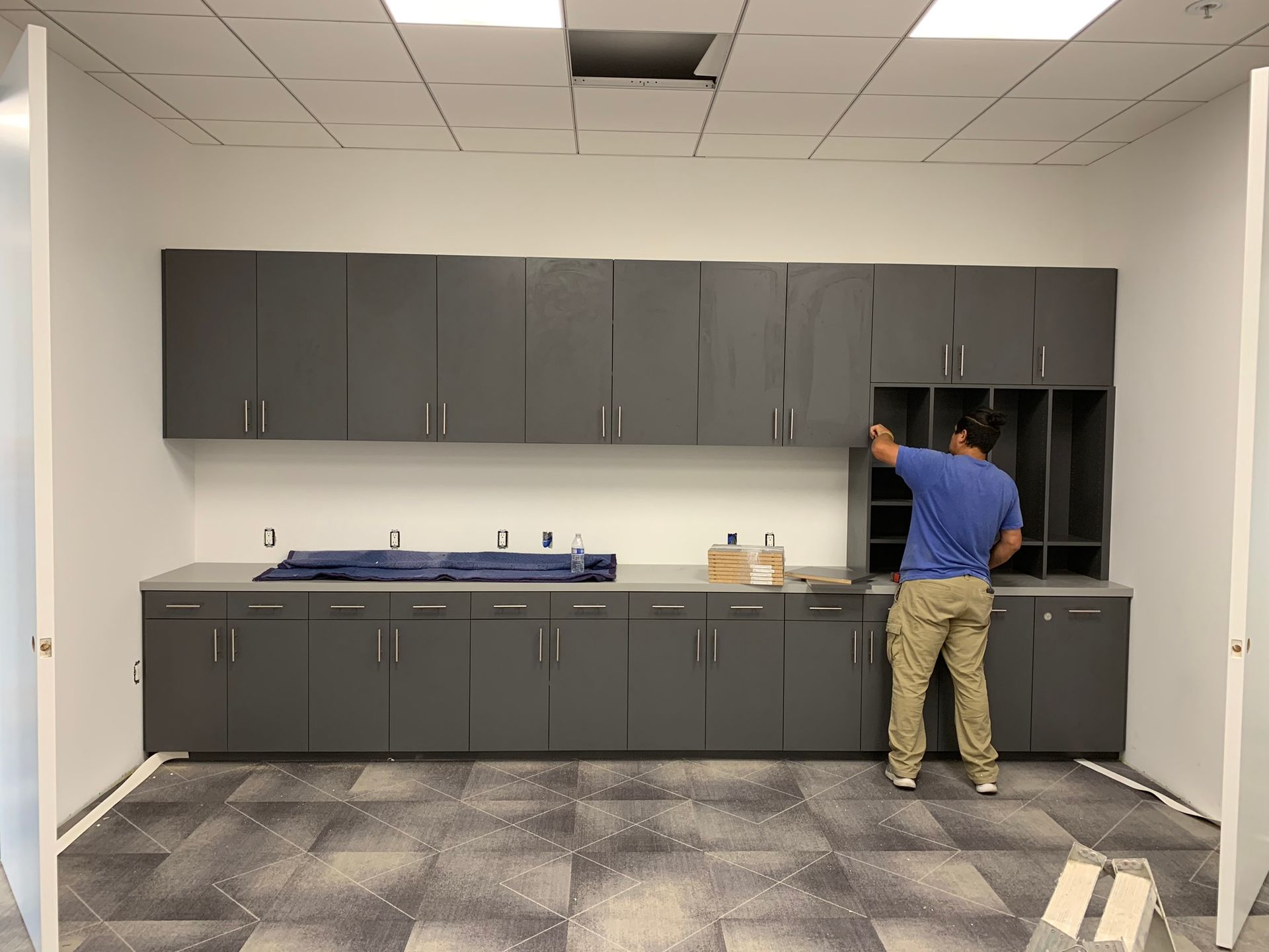 Man installing gray cabinets in a room with a gray countertop and open doorway, work in progress.