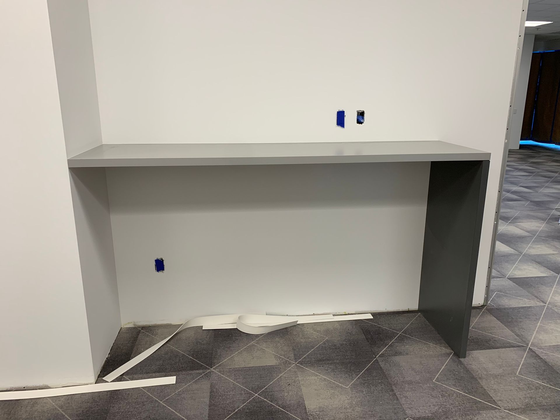 Gray desk against a white wall with blue outlet covers; floor with patterned carpet.