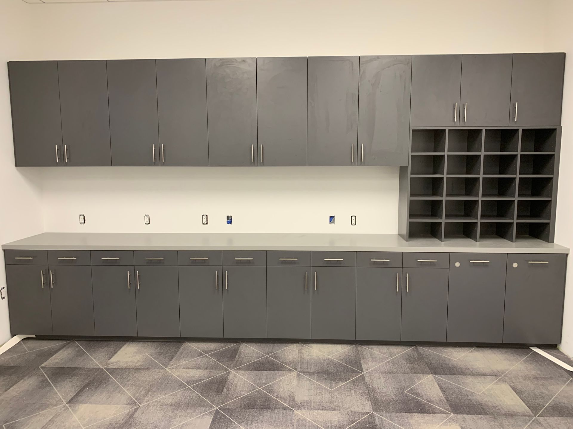 Gray cabinets with countertop and cubby storage, against a white wall and patterned flooring.