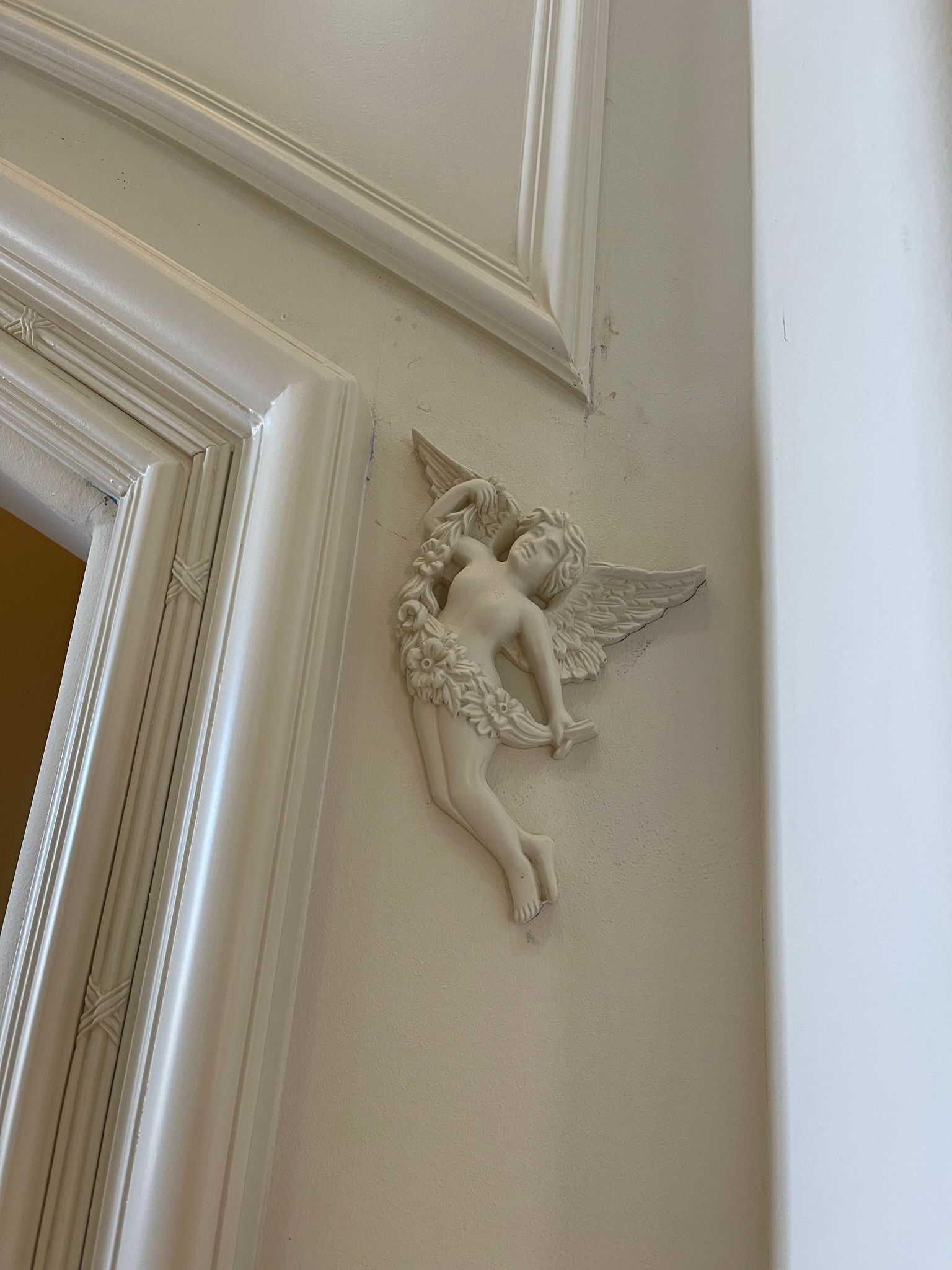White cherub sculpture with wings and floral garland on a white wall near a doorway.