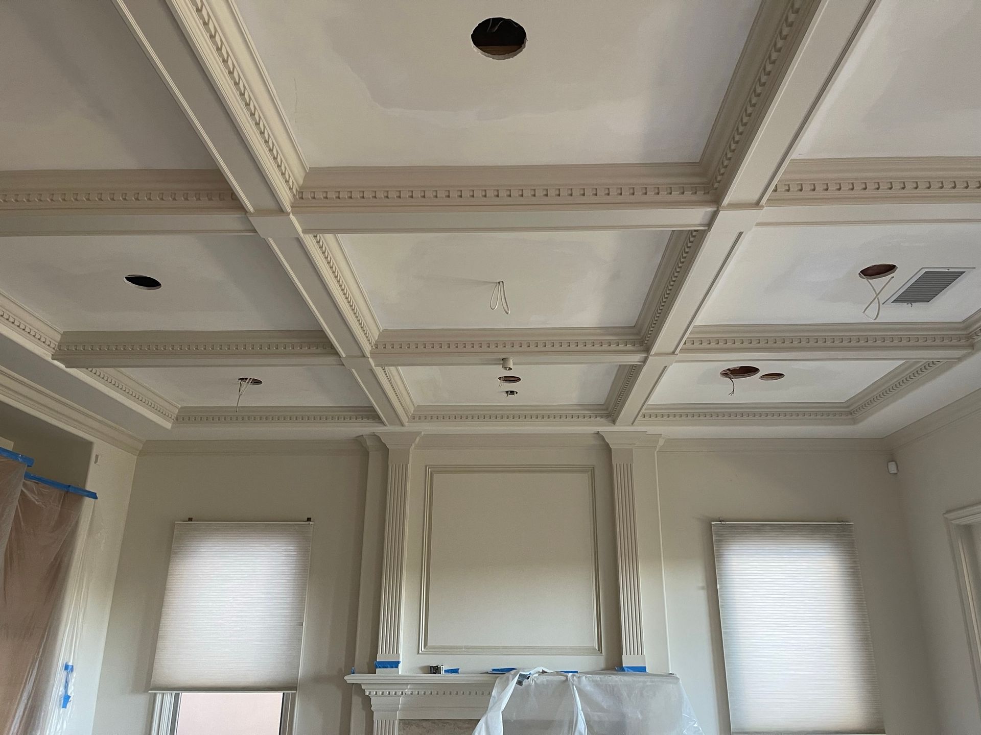 Ornate white coffered ceiling in a room with three windows and a fireplace. Construction in progress.