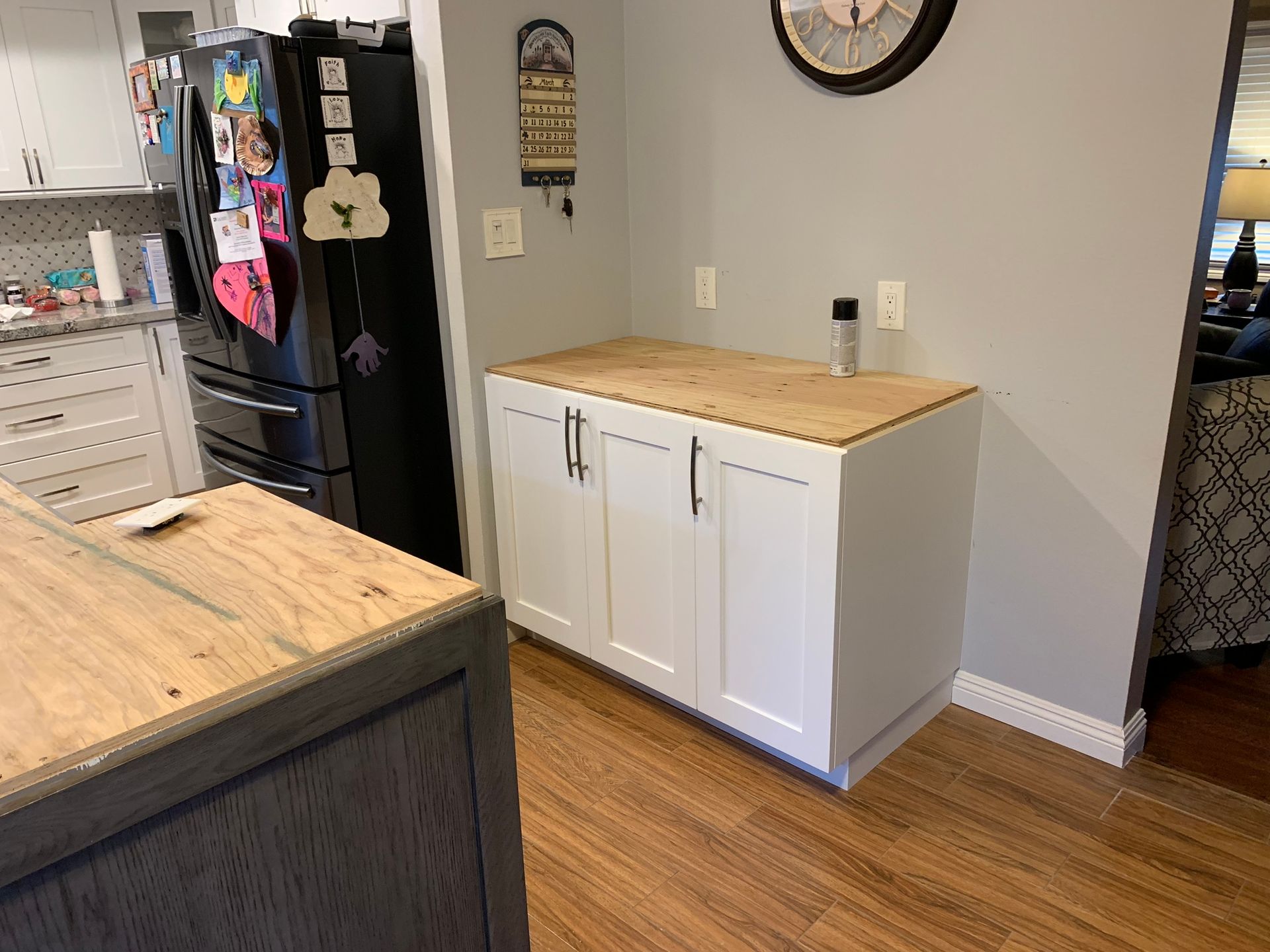 White kitchen cabinet with wooden countertop, against a gray wall, near a black refrigerator and wood floor.