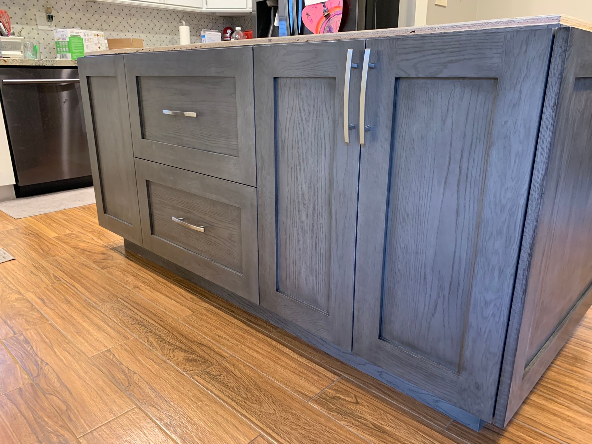 Gray kitchen island with cabinets and drawers, on a wooden floor, with a stainless steel appliance visible.