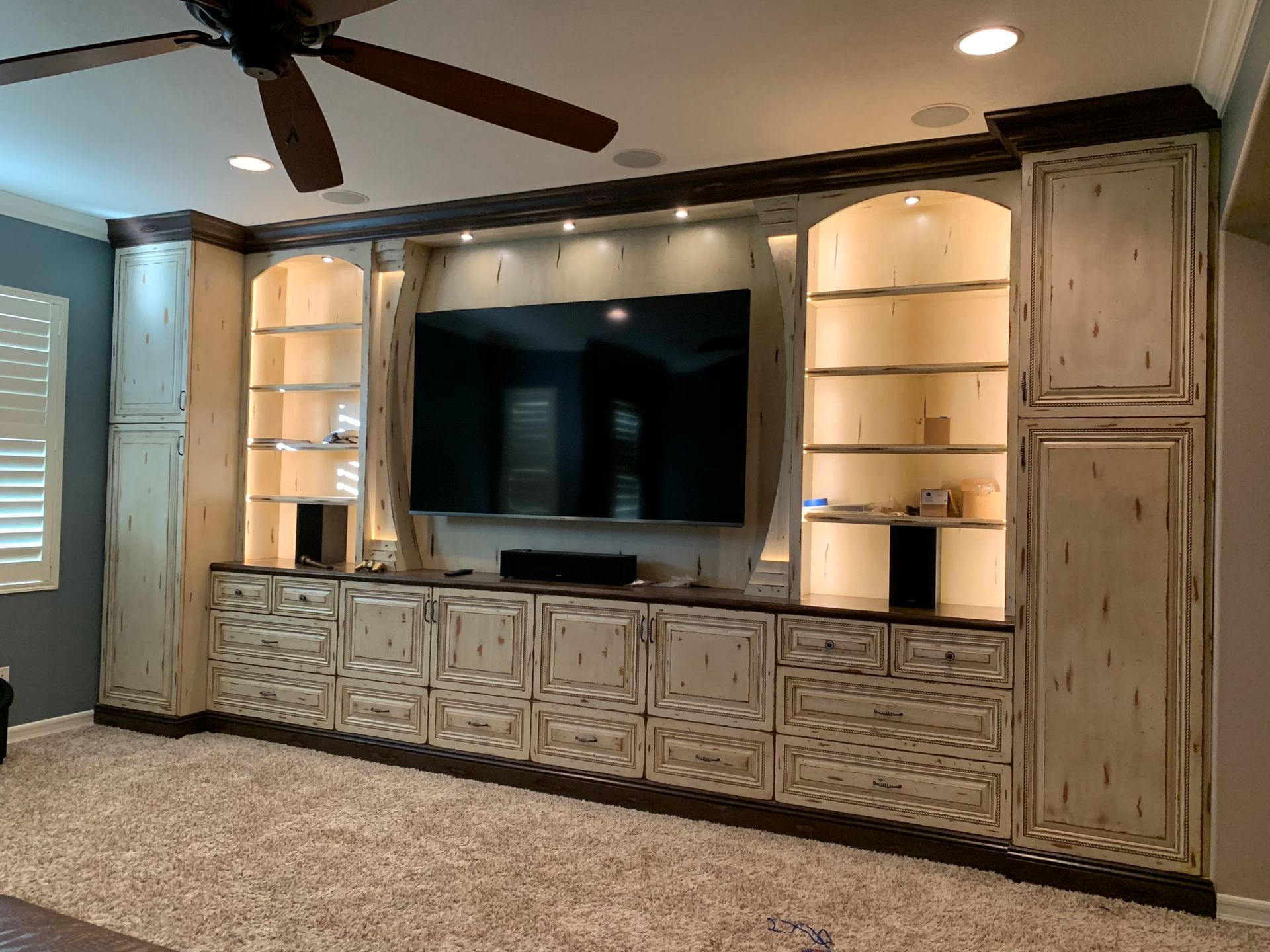 Built-in entertainment center with cream-colored cabinets, TV, and recessed lighting in a room with a ceiling fan.