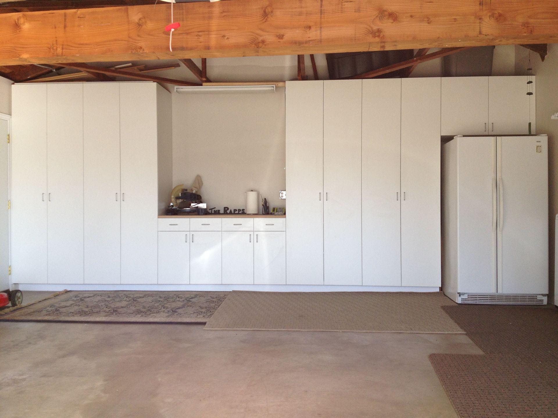 White cabinets and a refrigerator line a garage wall; an area rug lies on the floor.