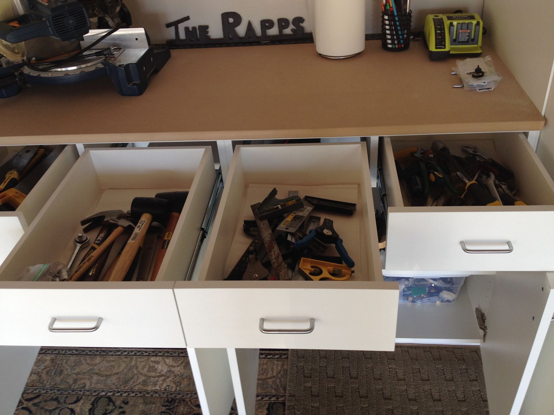 Open drawers in a white workshop cabinet with assorted tools and a workbench.