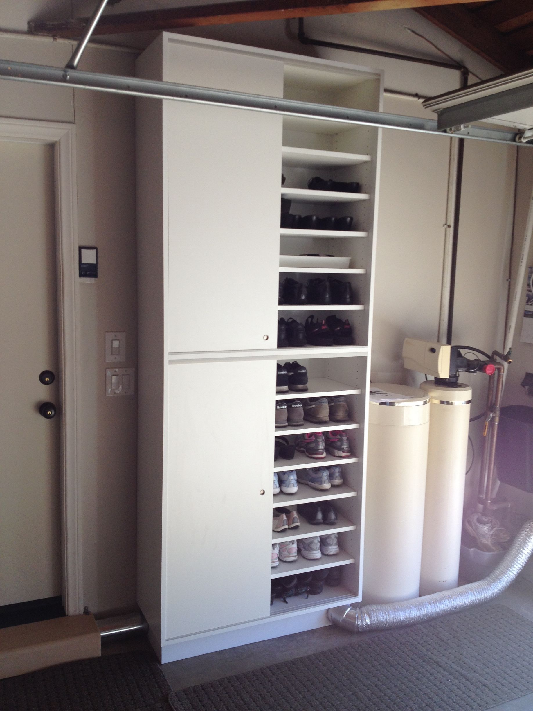 White shoe storage cabinet in a garage next to water filtration system, with closed doors and open shelves displaying shoes.