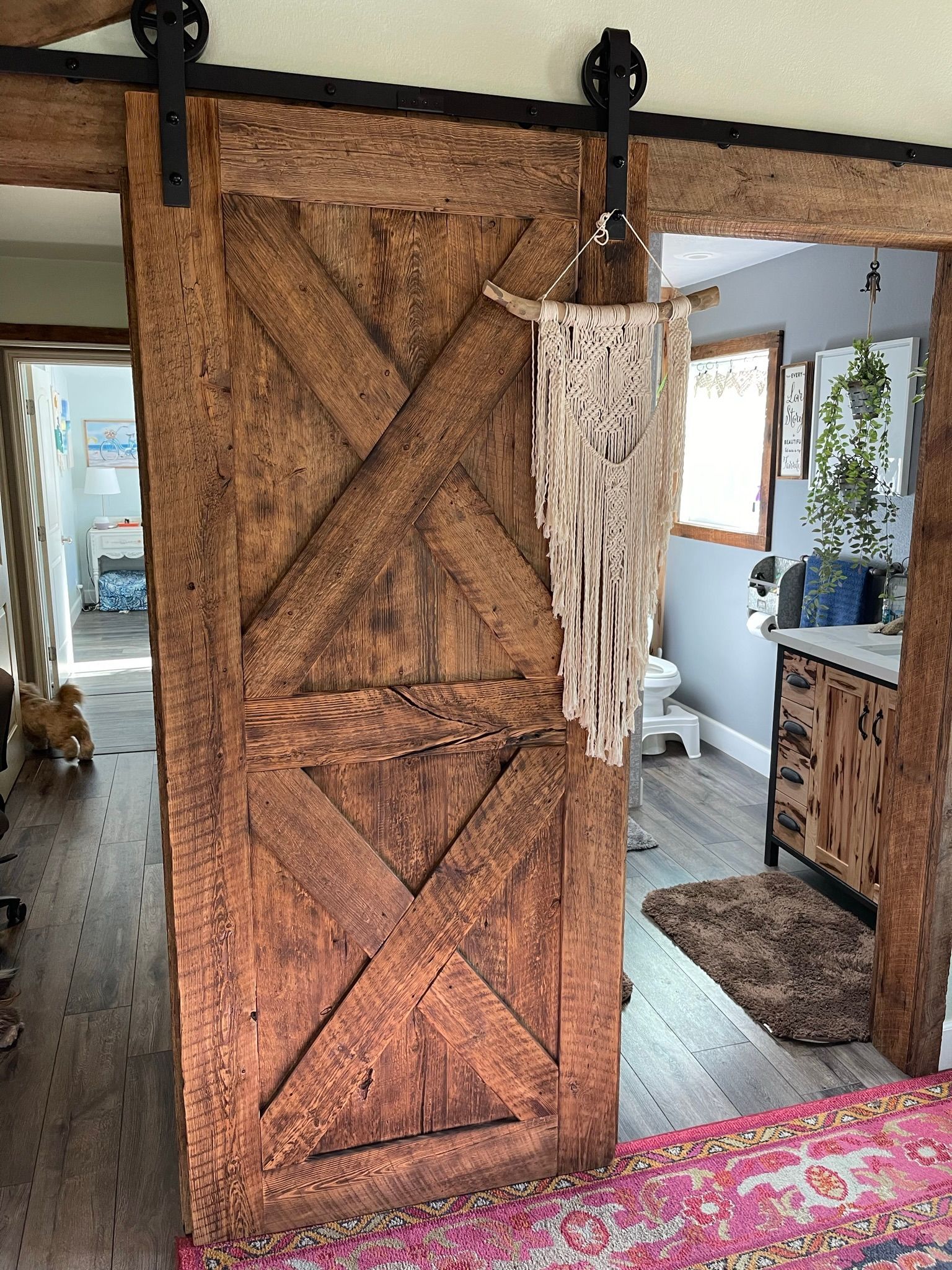 Rustic wooden barn door partially open, revealing a room with a decorative macrame hanging, rug, and other decor.