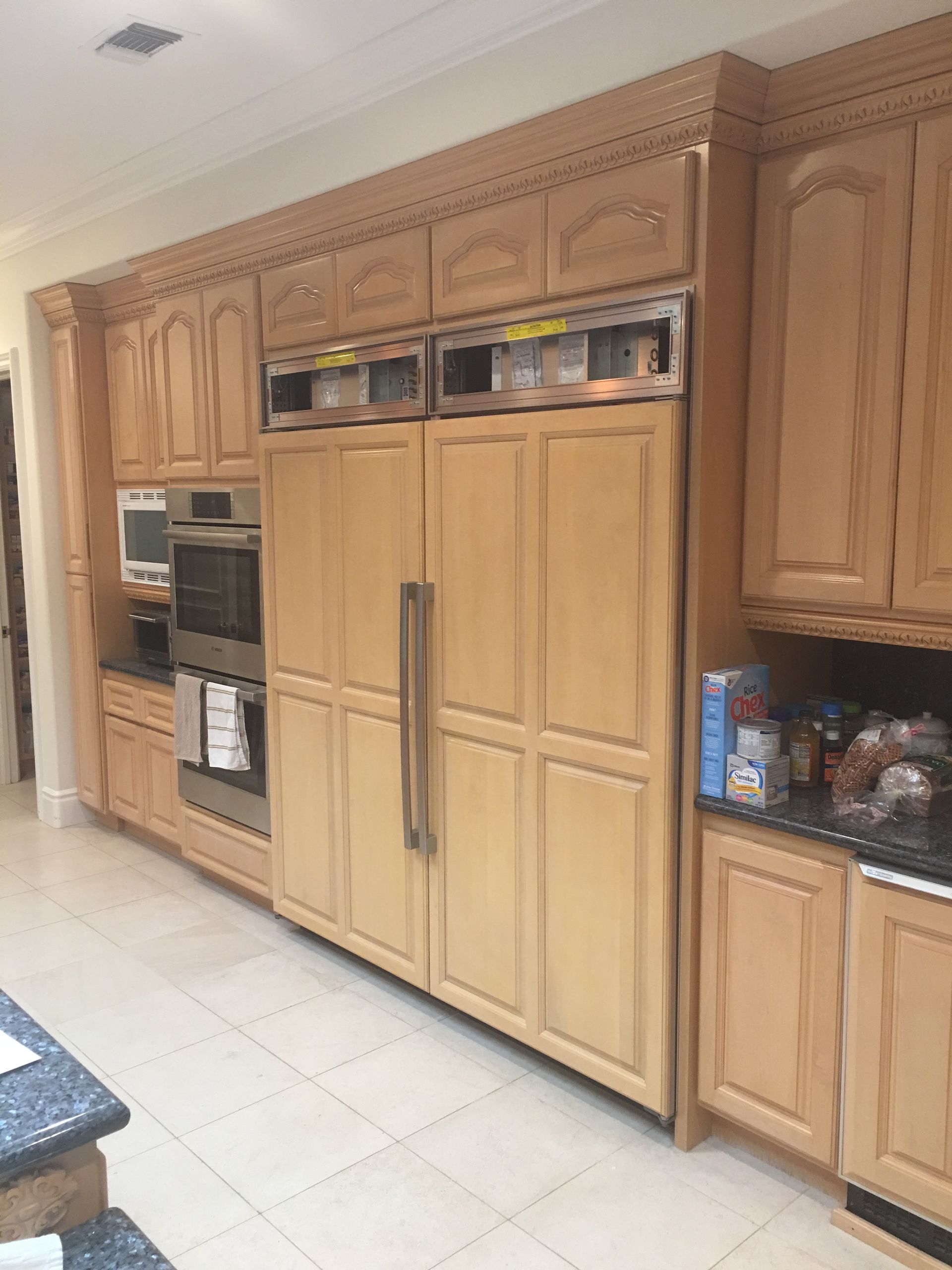 Kitchen cabinets and built-in refrigerator with a double oven. Light wood cabinetry and white tiled floor.