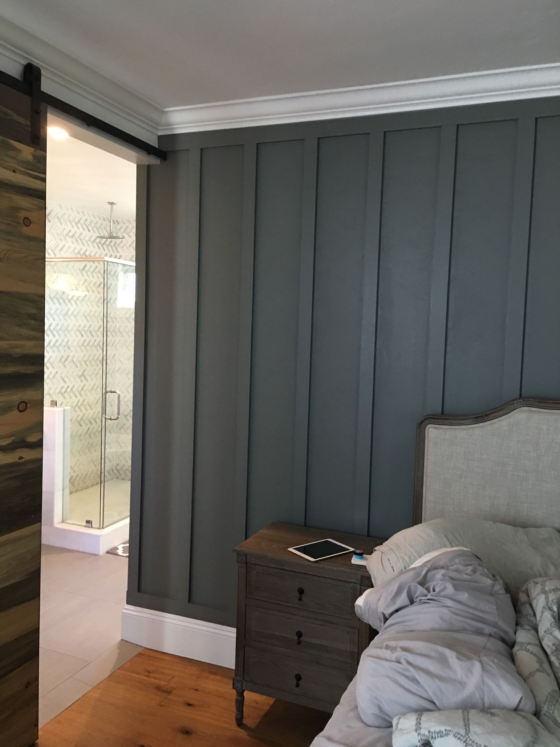 Bedroom with gray paneled wall, a wooden nightstand, and a sliding barn door leading to a shower.
