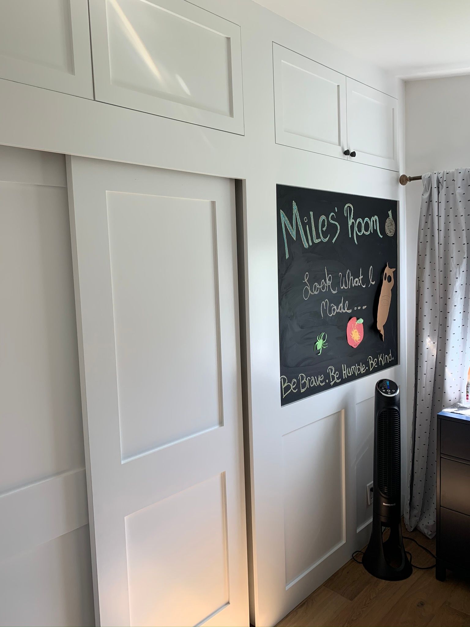 White built-in closet with sliding door, chalkboard sign, and fan against a white paneled wall.