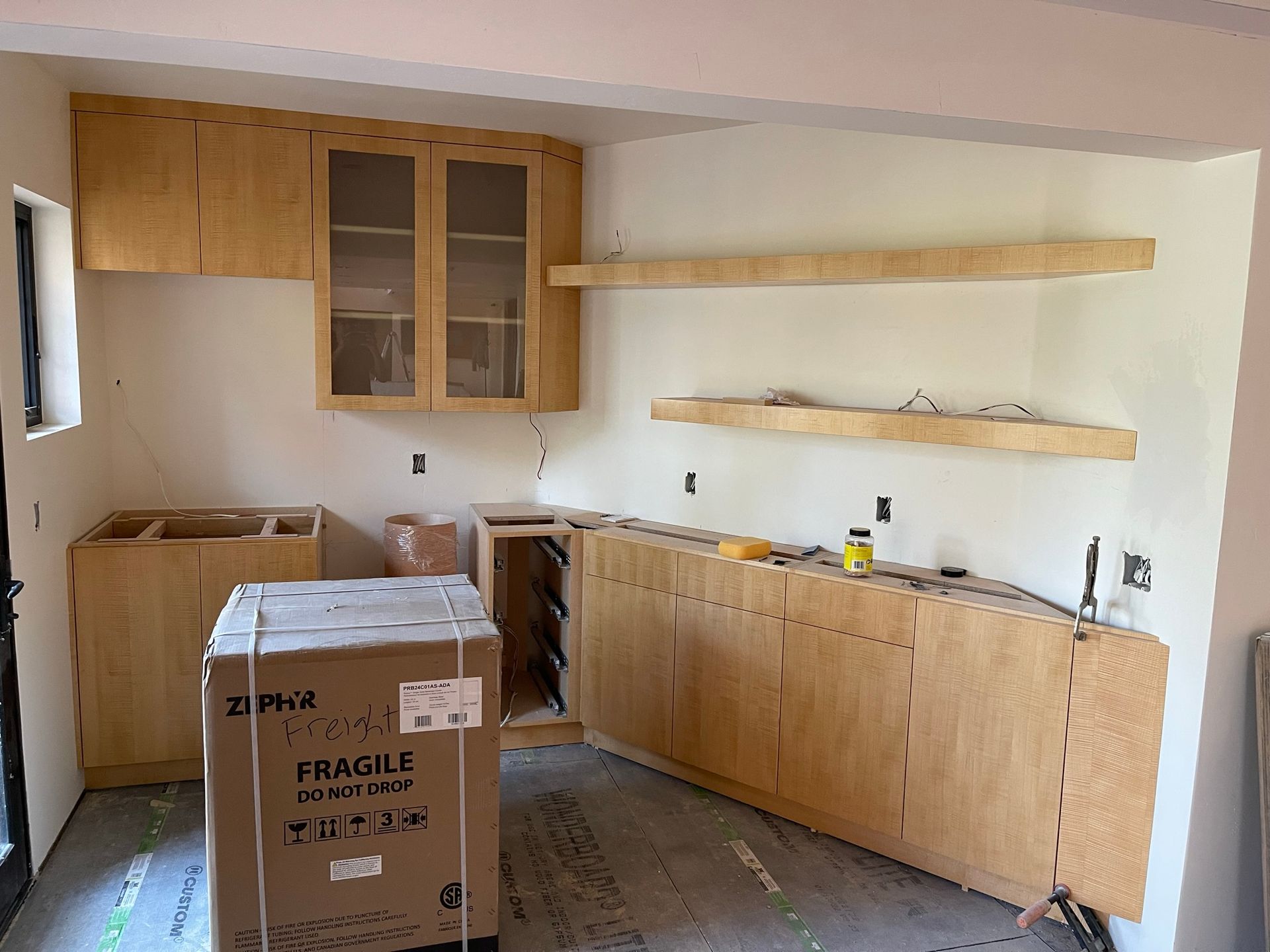 Kitchen under construction with light wood cabinets and shelves. Unfinished walls and appliances.