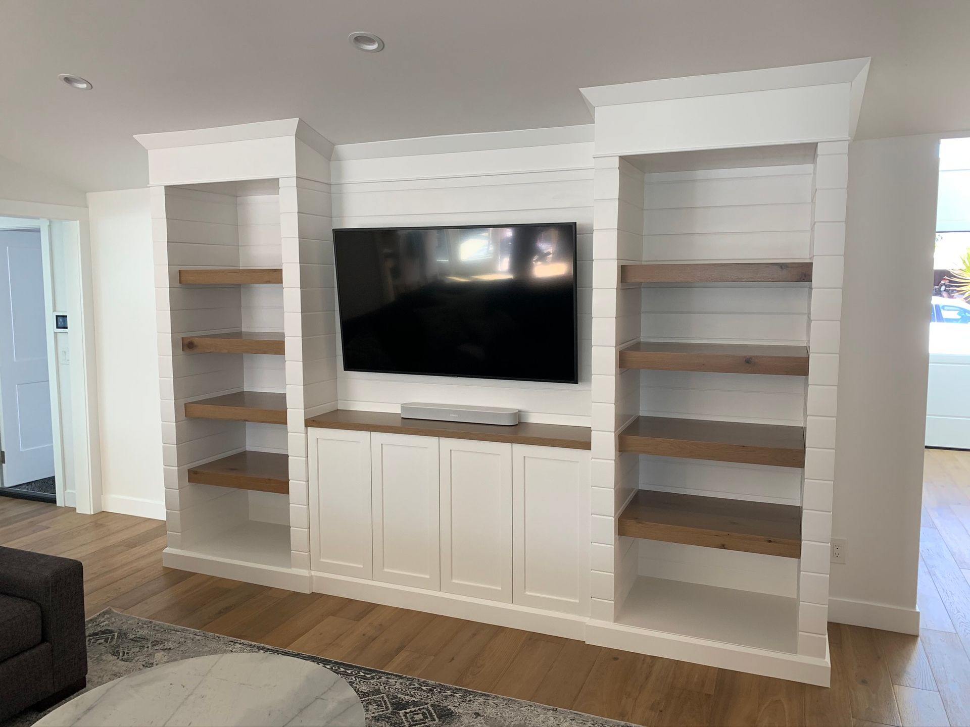 White built-in entertainment center with a TV, shelving, and cabinets against a white wall with wooden floors.