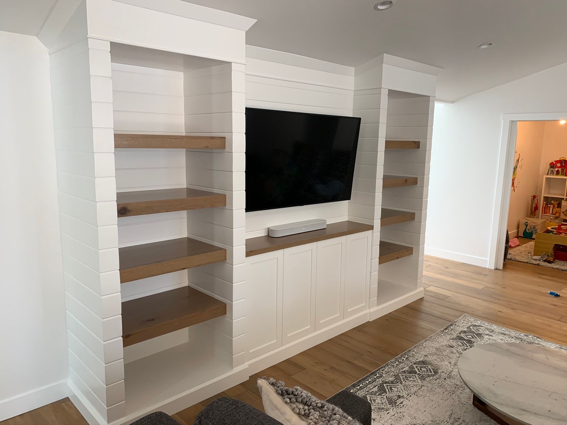 White built-in shelves with wood shelves and cabinetry framing a mounted TV; doorway in the background.