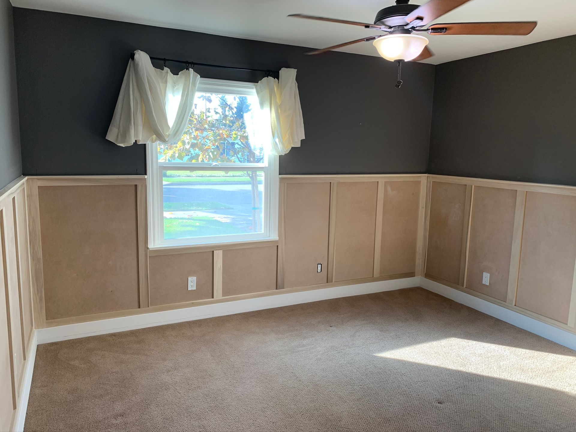 Empty bedroom with gray walls, beige wainscoting, a window, tan carpet, and a ceiling fan.
